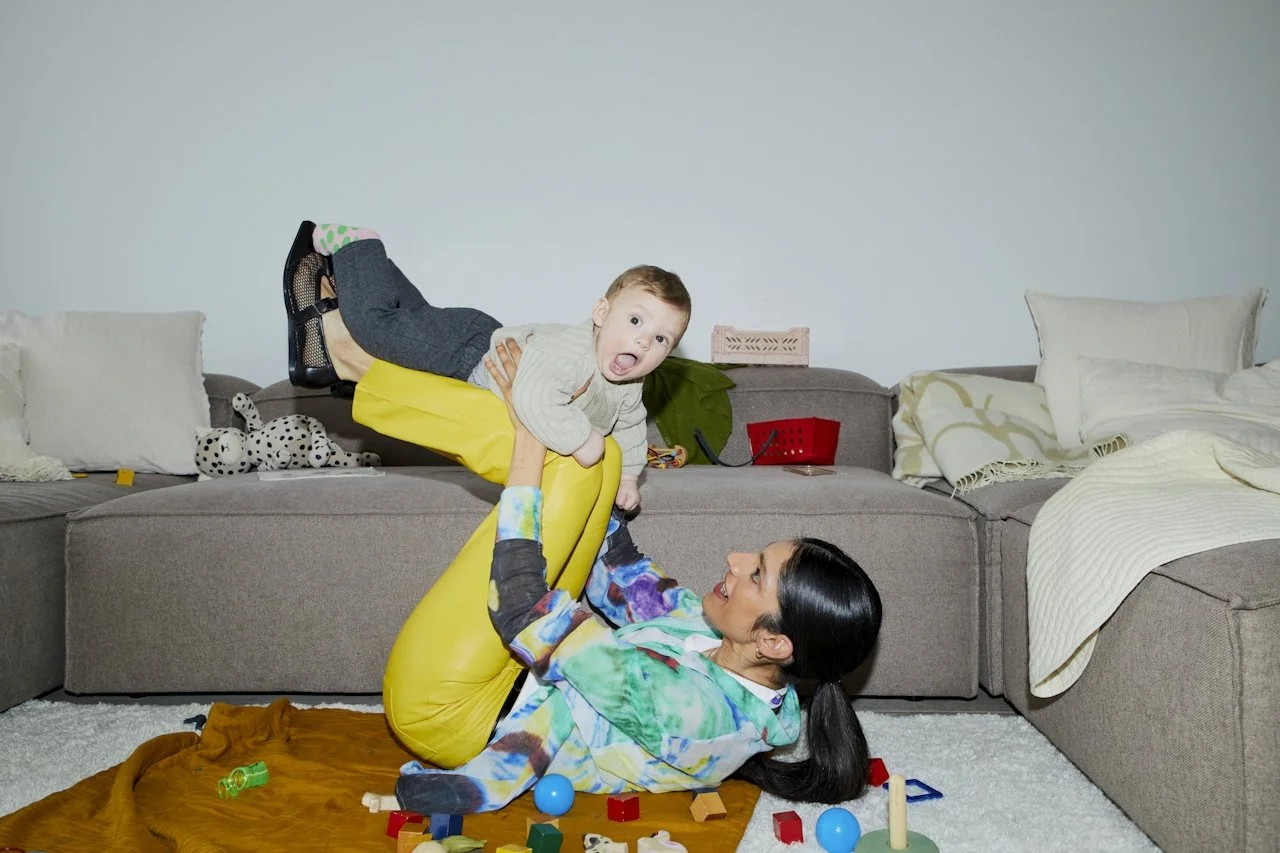 Woman lying on the floor playing with a young boy who is balancing on an inflatable yellow armrest of a couch, with toys scattered around.