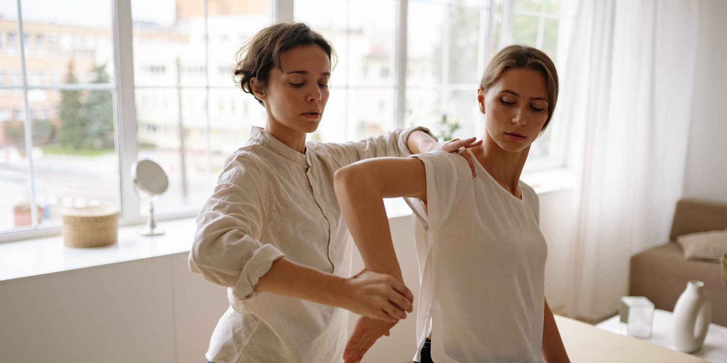 A woman receives a shoulder massage from a massage therapist in a bright room with large windows and white curtains.