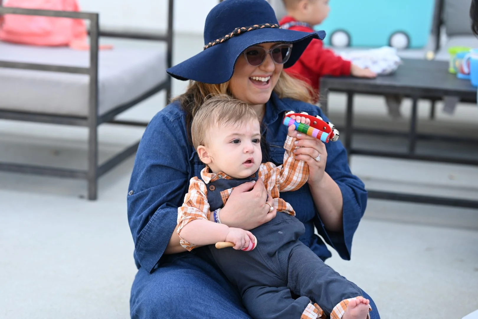 A woman wearing a blue wide-brimmed hat and sunglasses holding a young boy, who is looking surprised. The woman is smiling and sitting on the ground, with a child in the background at a table.