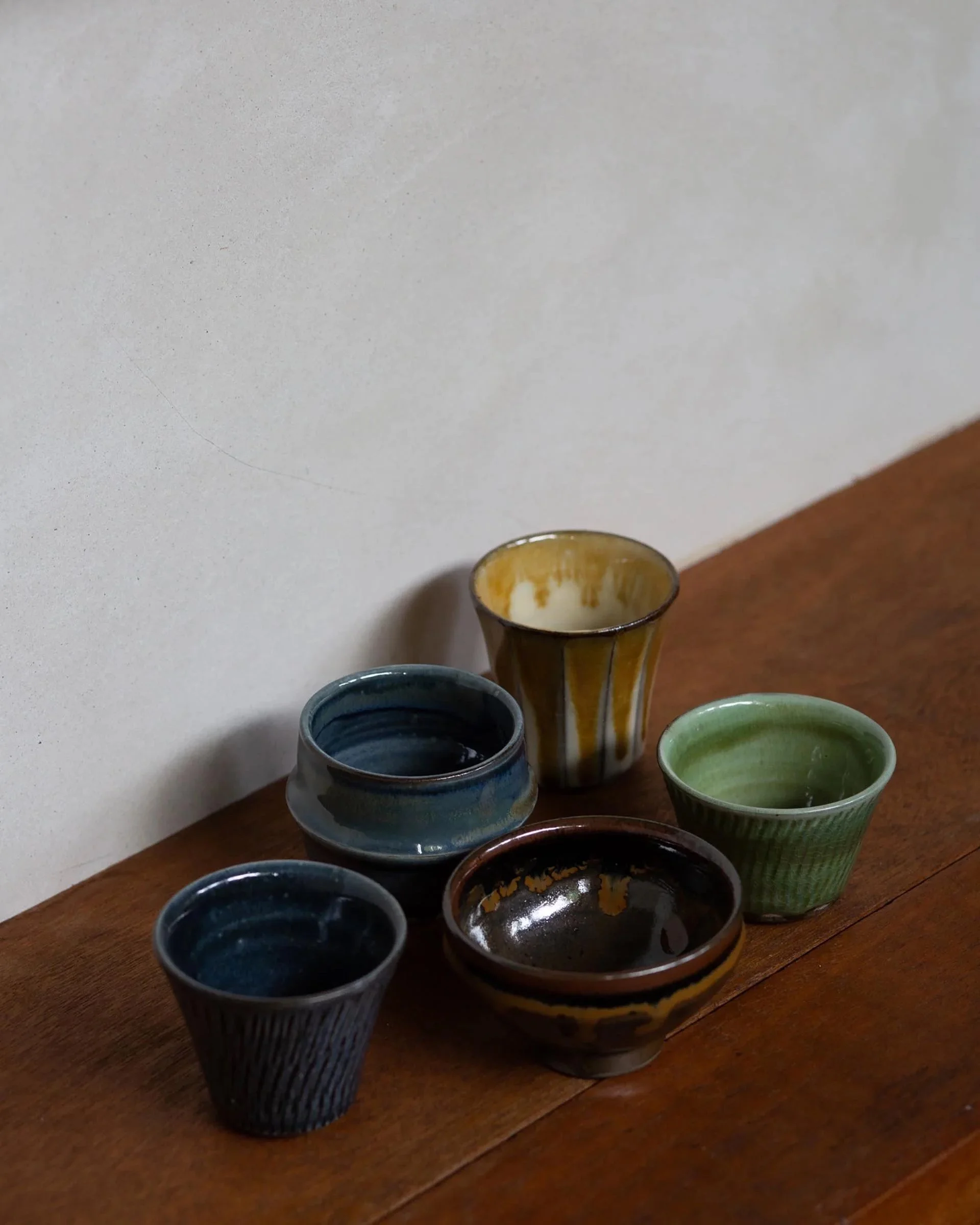 Assorted hand-glazed sake cups in earthy tones – green, blue, brown, yellow – grouped on a wooden surface. Each cup displays individual glaze variation.