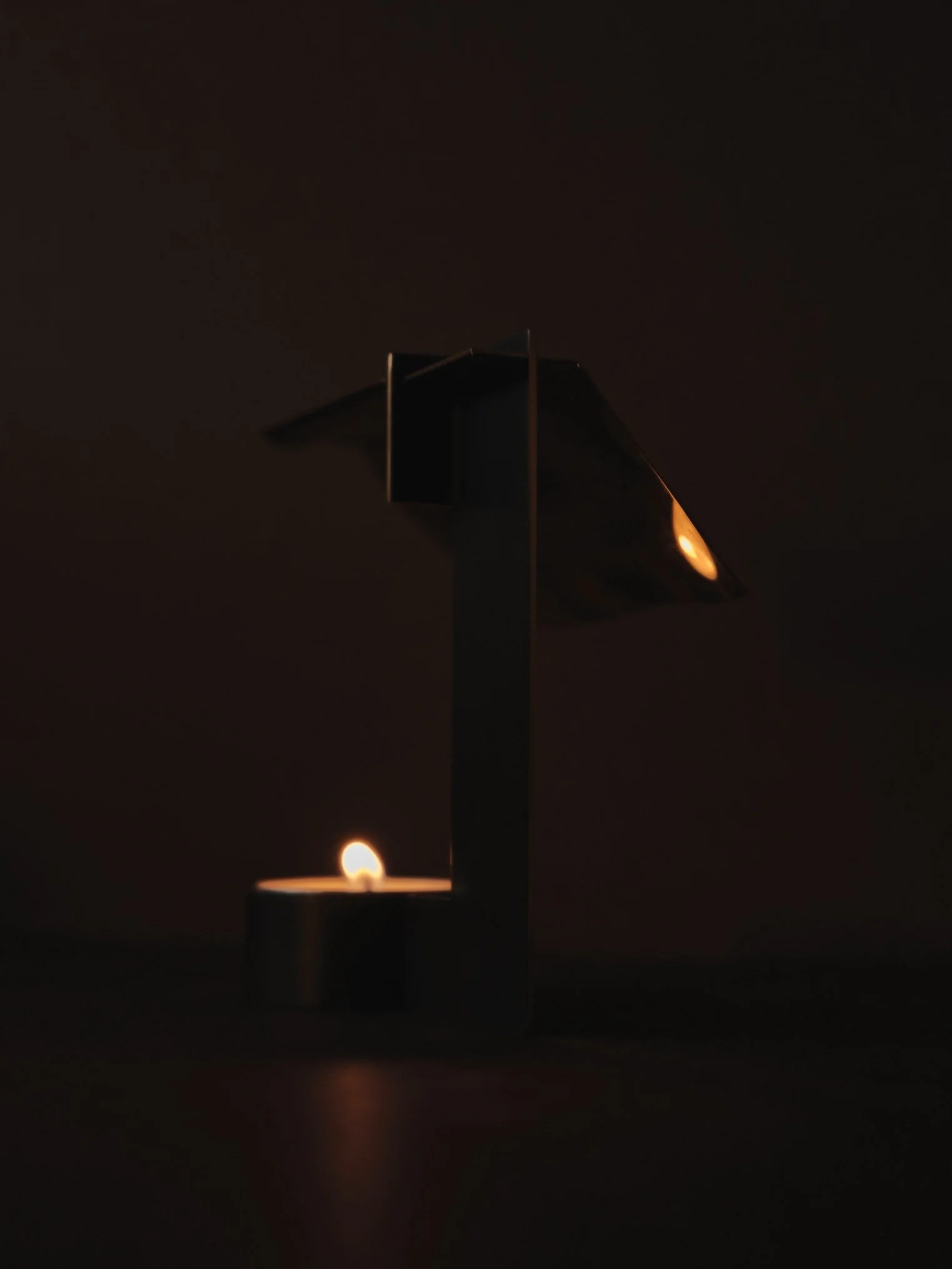 Designed by Chialing Chang. Moody minimalist close-up of a modern black metal candle holder with a warm glowing tealight, set against a dark background, creating an intimate, sculptural home décor lighting scene.