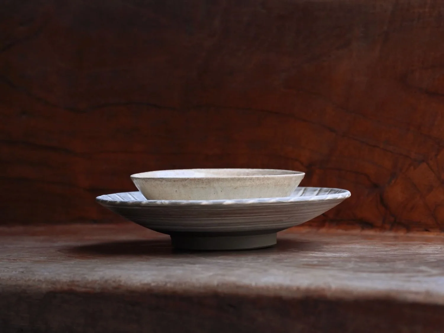 Side profile of a handmade ceramic plate and cup set with a gently curved rim and subtle crackled glaze, set against a wooden background