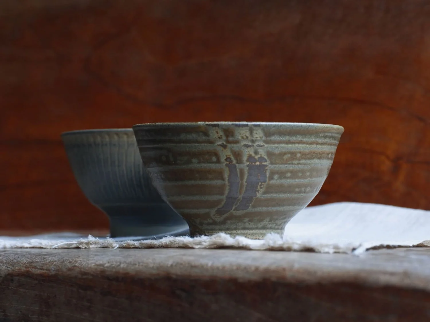 Two Onta ware pottery bowls with vertical slip-glaze designs, side view, resting on white fabric against a wood backdrop