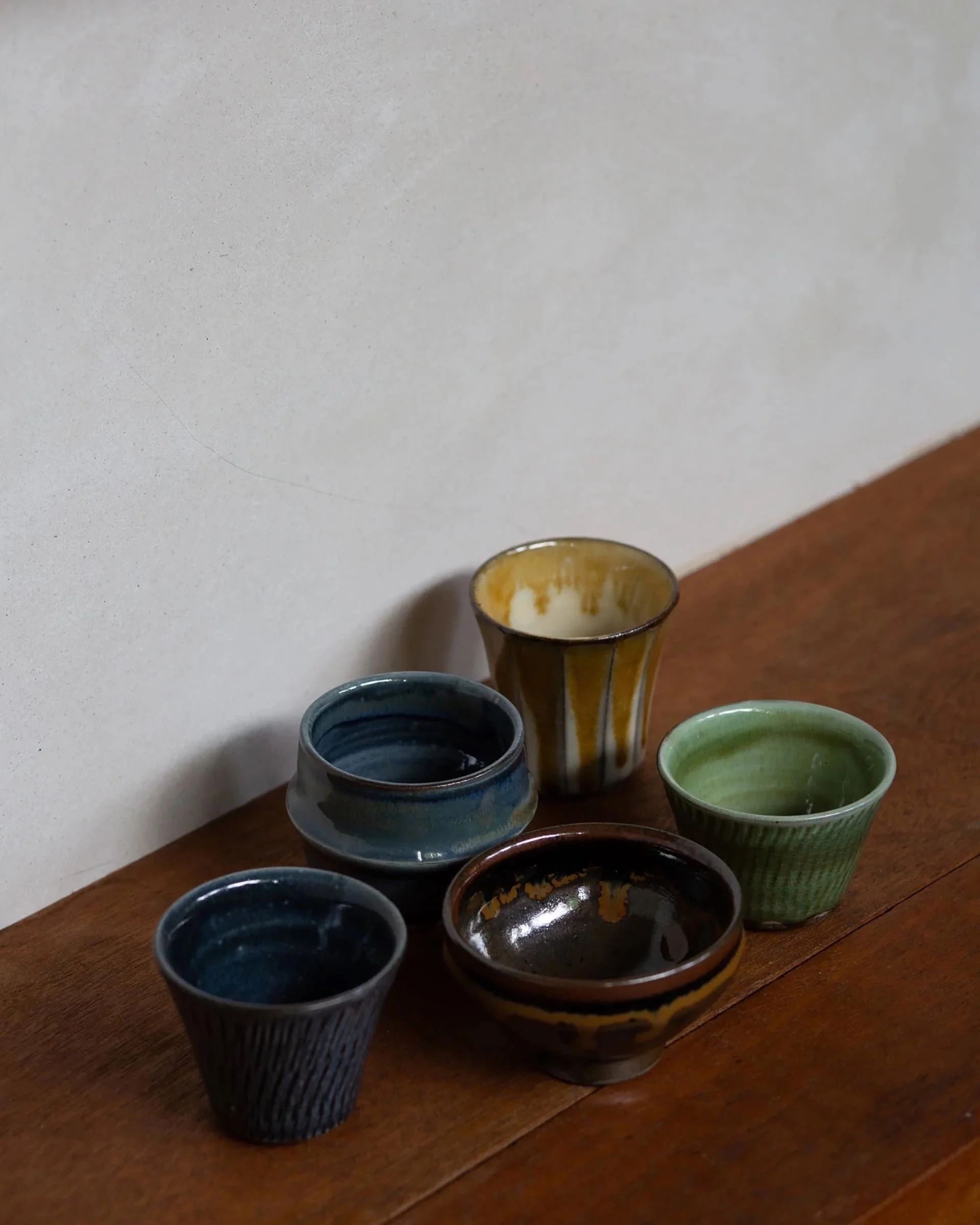 Assorted hand-glazed sake cups in earthy tones – green, blue, brown, yellow – grouped on a wooden surface. Each cup displays individual glaze variation.