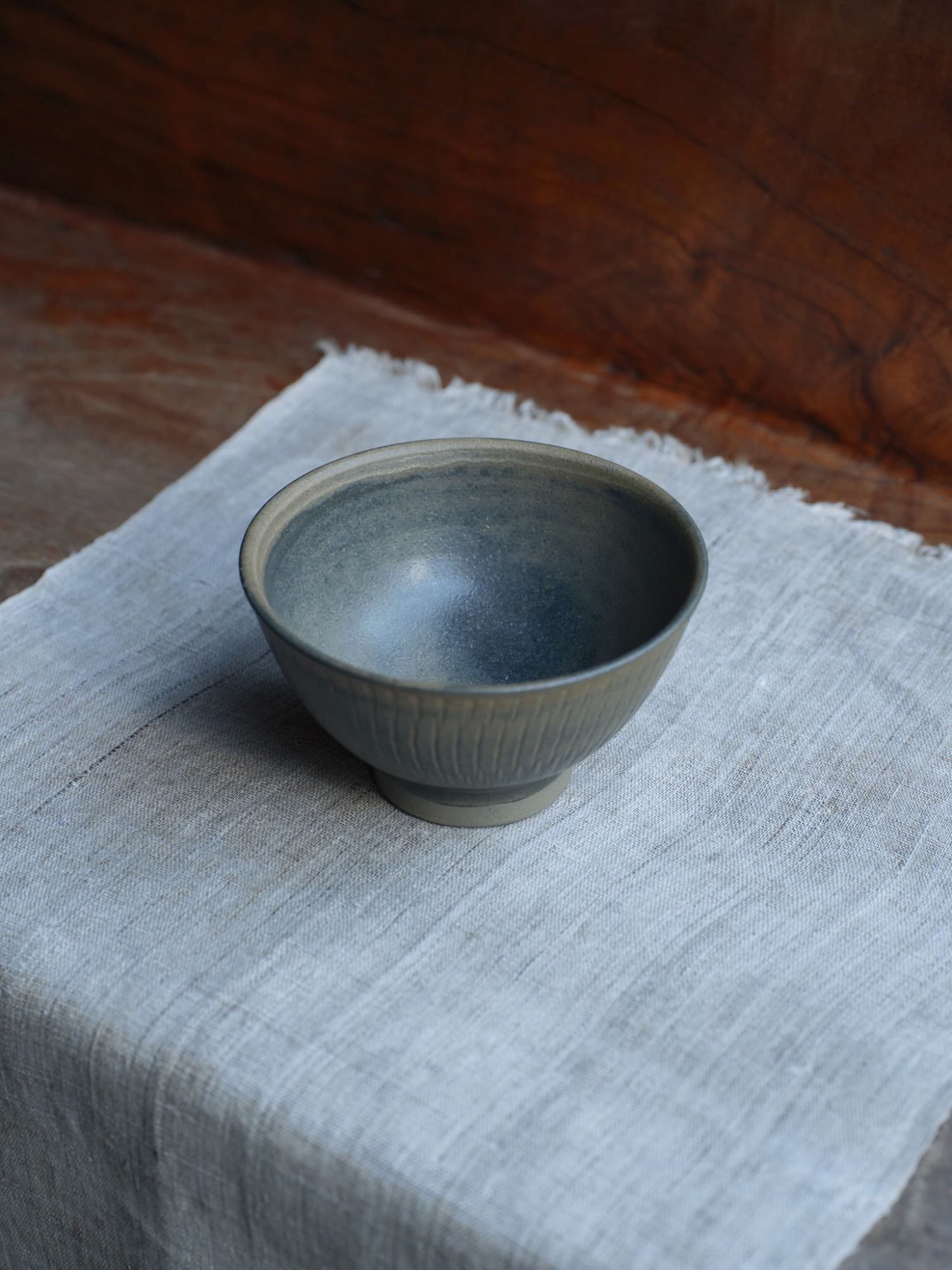 Single handmade ceramic bowl with fine linear texture and deep glaze, positioned on a soft linen mat