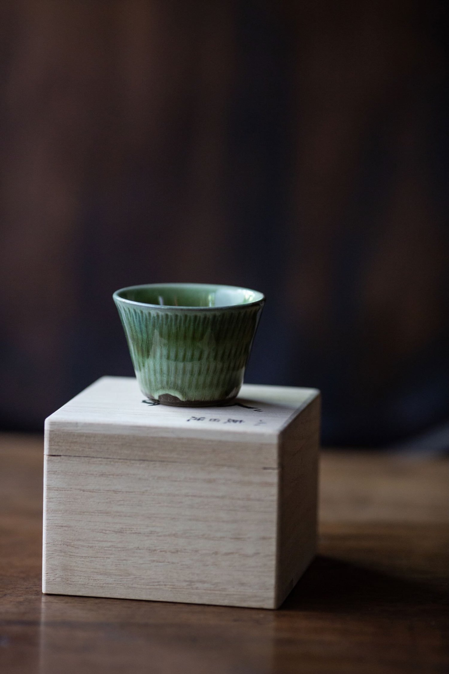 Green sake cup with vertical glaze pattern, resting on a wooden box with Japanese calligraphy.