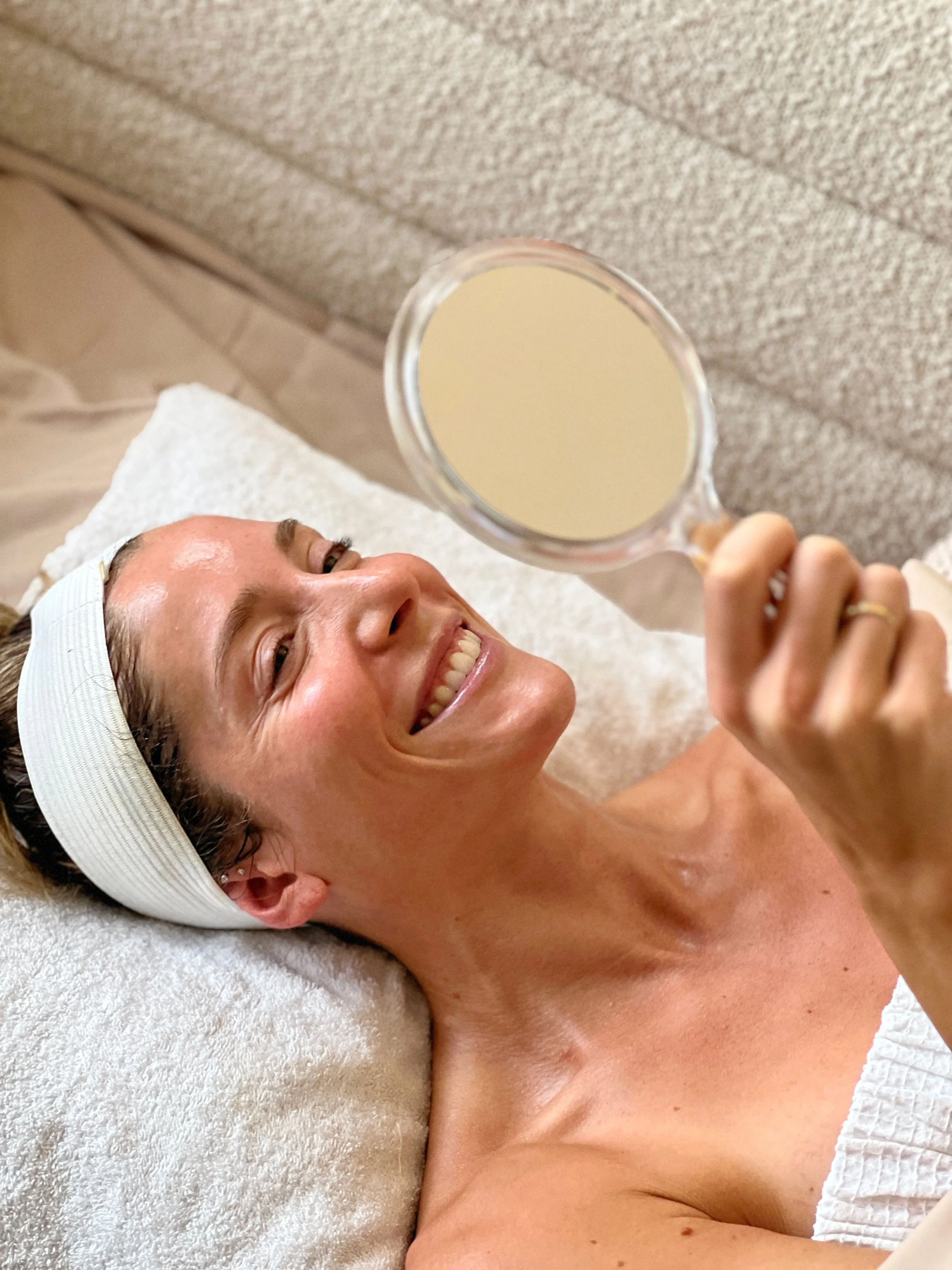 A woman lying on a spa bed with a headband, smiling and holding a small mirror in front of her face.