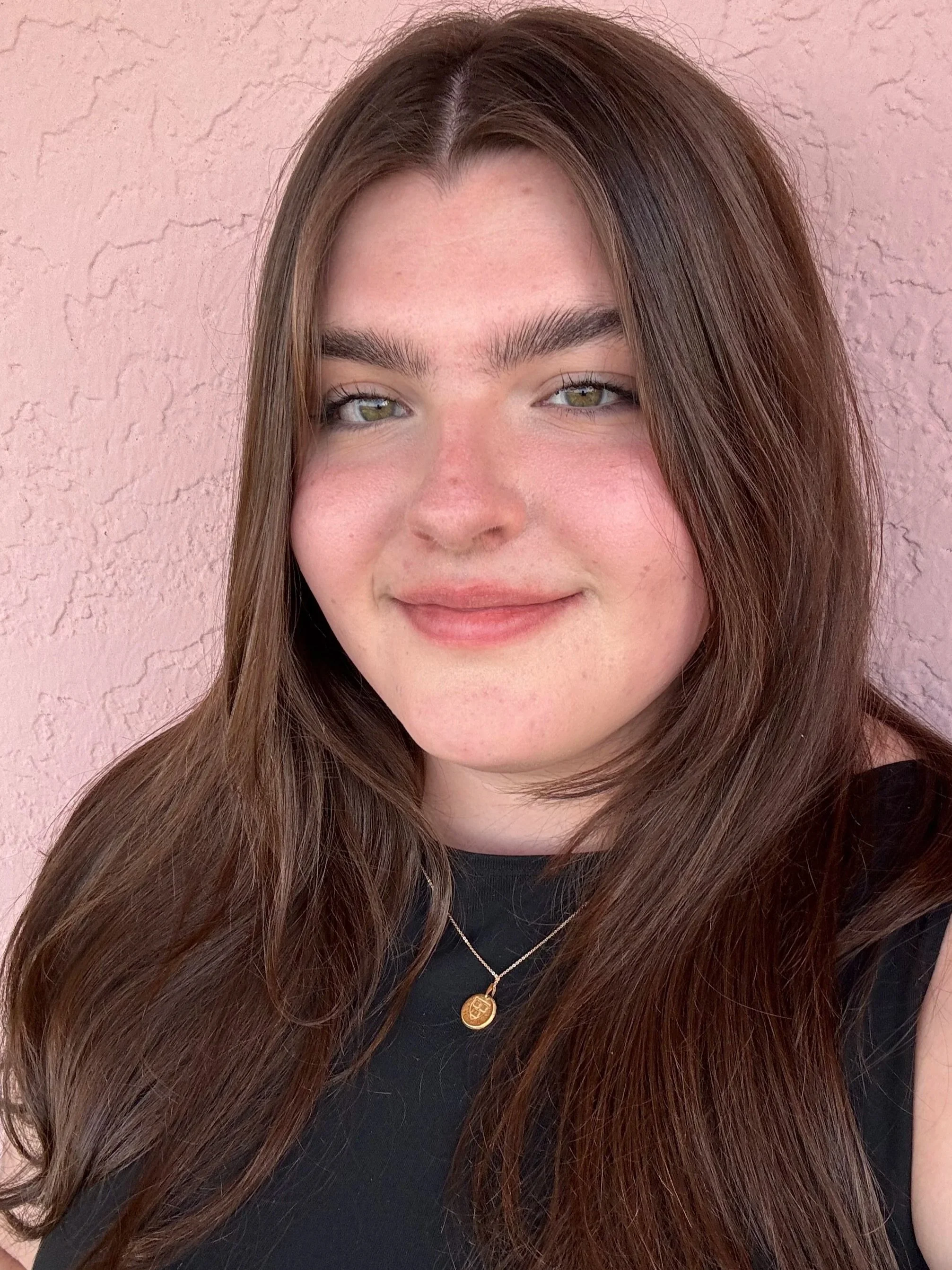 Close-up of a young woman with long, wavy brown hair, wearing a black shirt and a gold necklace with a pendant, standing against a pink textured wall.