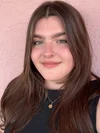 Close-up of a young woman with long, wavy brown hair, wearing a black shirt and a gold necklace with a pendant, standing against a pink textured wall.