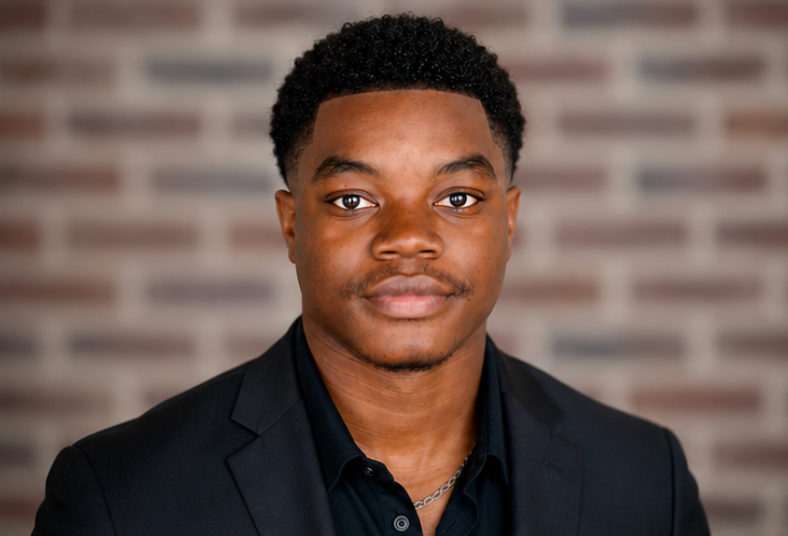 Portrait of a young African American man in a black suit and black shirt, standing in front of a brick wall background.