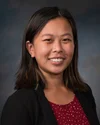 Close-up portrait of an Asian woman smiling, wearing a black blazer over a red top with white polka dots, against a dark gray background.