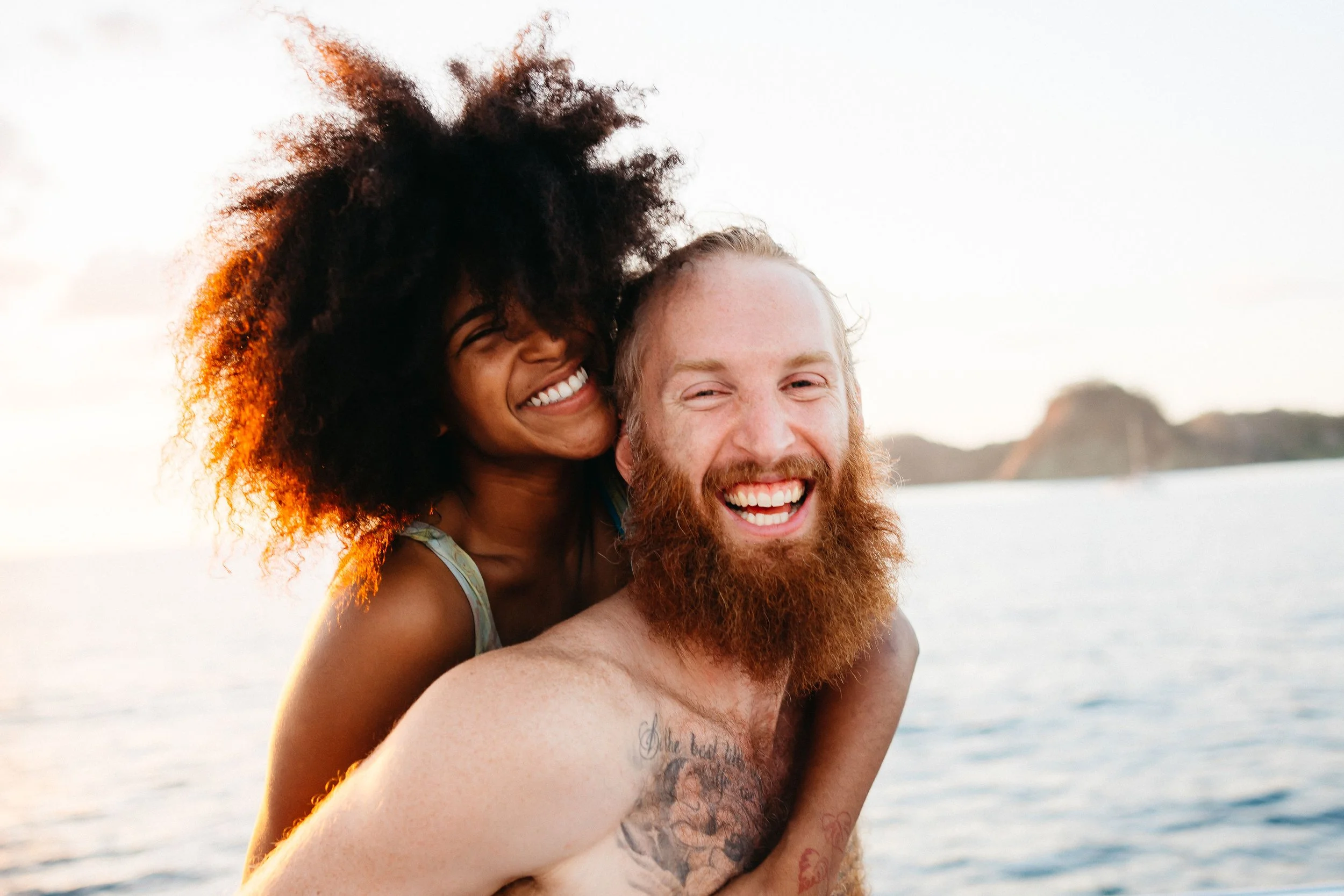 A joyful couple, a woman with curly hair and a man with a beard, smiling and playing by the water during sunset at the beach.