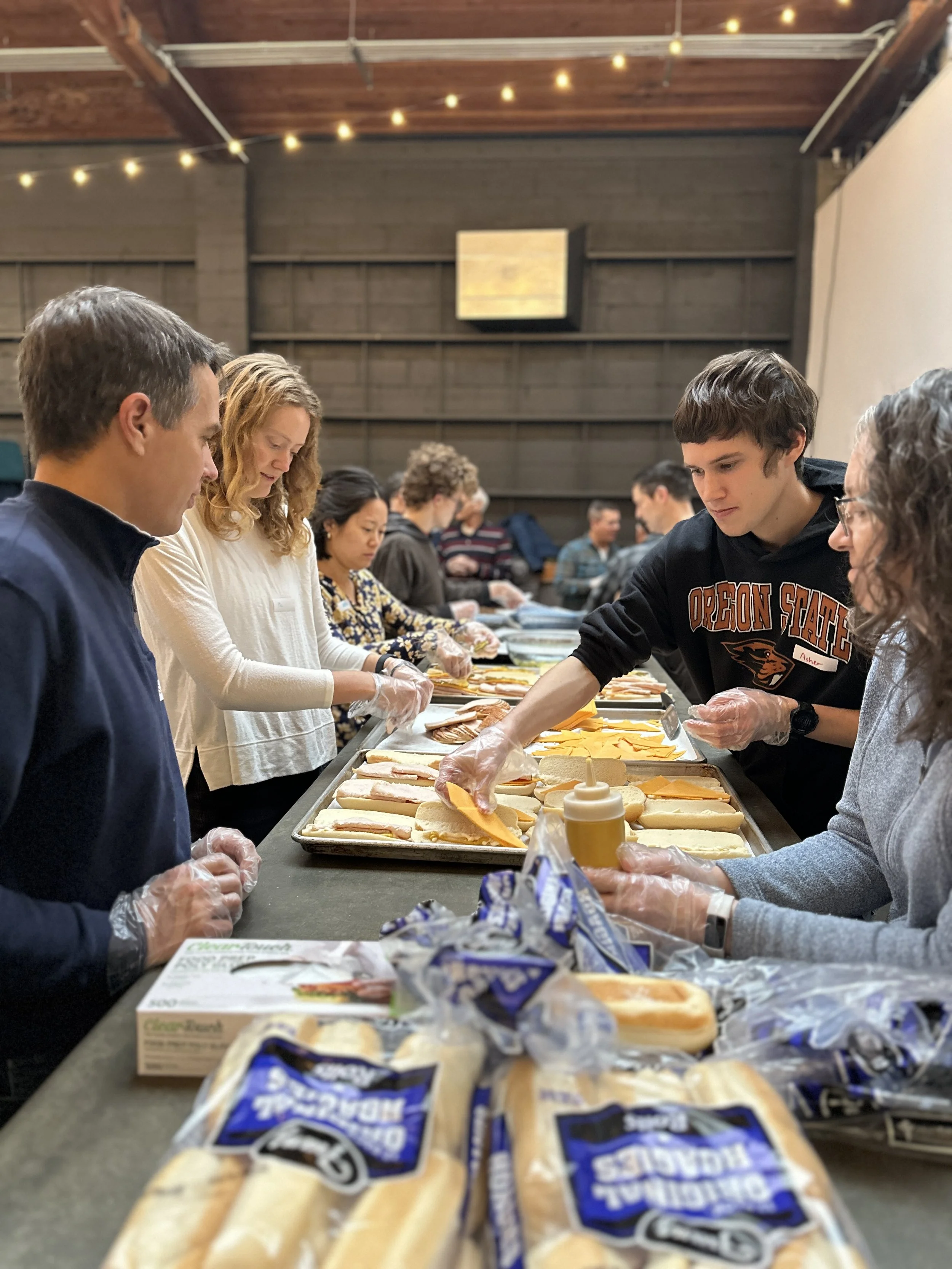 People preparing sandwiches at a long table, with ingredients like cheese, bread, and cold cuts, in an indoor setting with string lights.