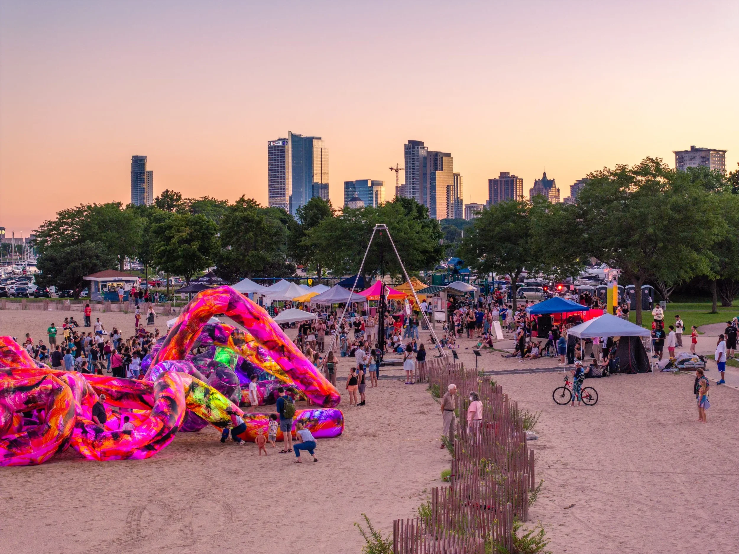 A lively outdoor event at sunset with colorful inflatable art sculptures in the foreground, a crowd of people, tents, and trees, with a city skyline of tall buildings in the background.