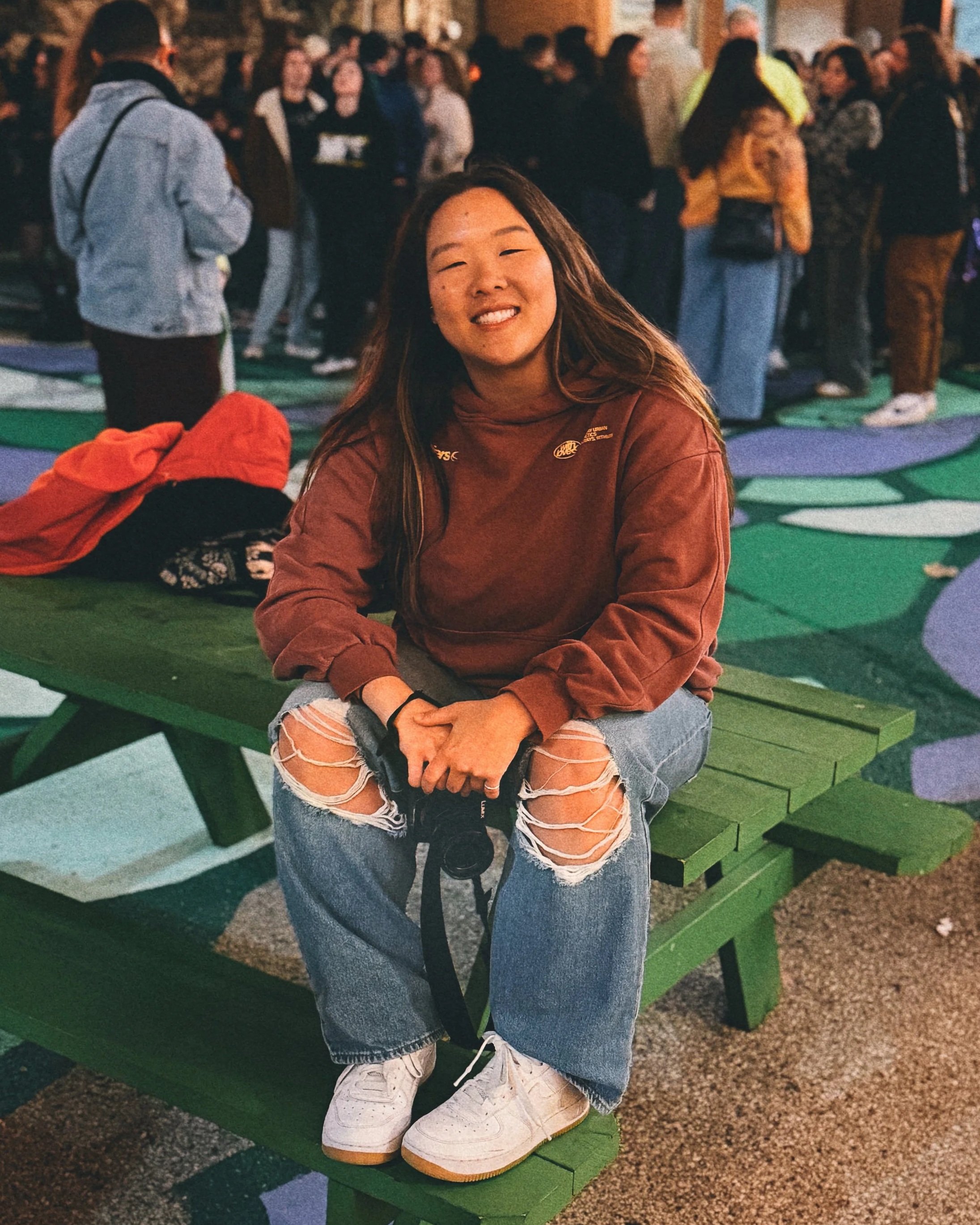 Melaina Zetley sitting on a green picnic table holding a camera with a group of people behind her.