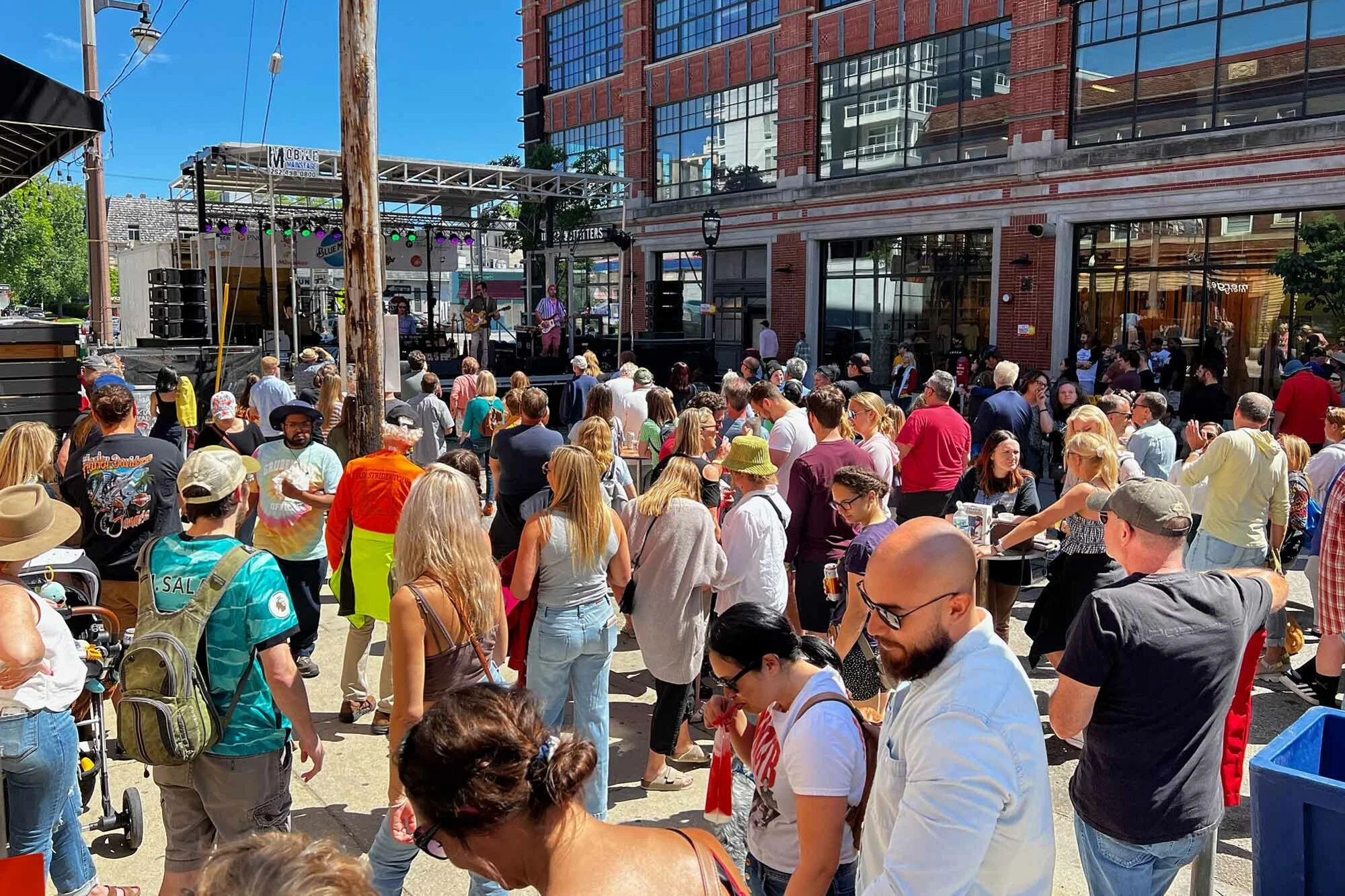 A crowd of people gathered outdoors on a sunny day, watching a live band perform on a stage in front of a large brick building with glass windows.