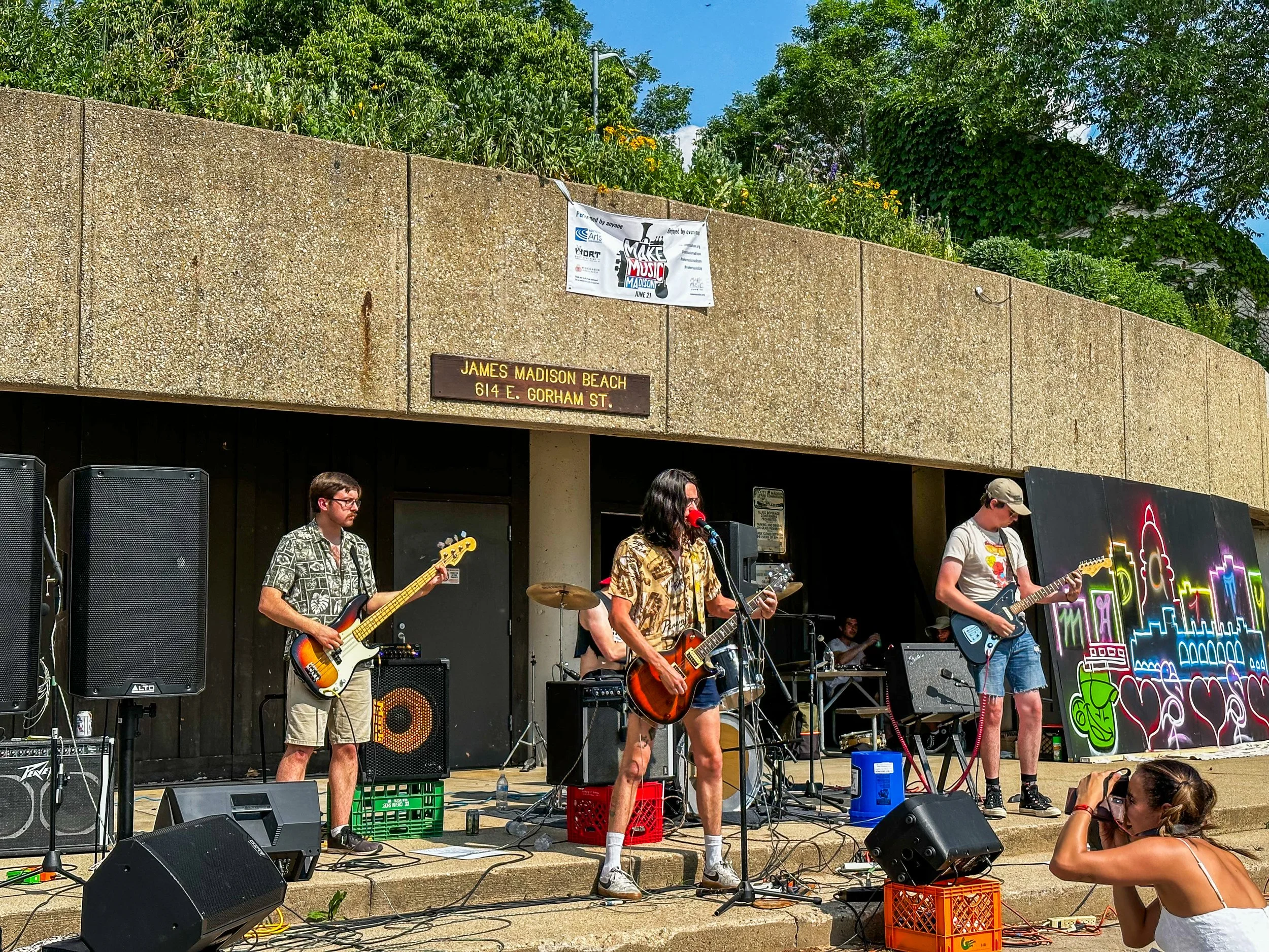 A group of musicians performing live on an outdoor stage with colorful city skyline artwork and a sign reading James Madison Beach, 614 E. Gorham St. People are playing guitars and singing, with speakers and sound equipment surrounding them.