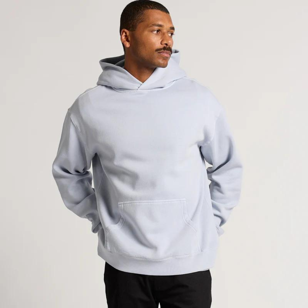 Man wearing a light gray hooded sweatshirt with kangaroo pocket, standing with hands in pockets, looking to the side, against a plain light background.