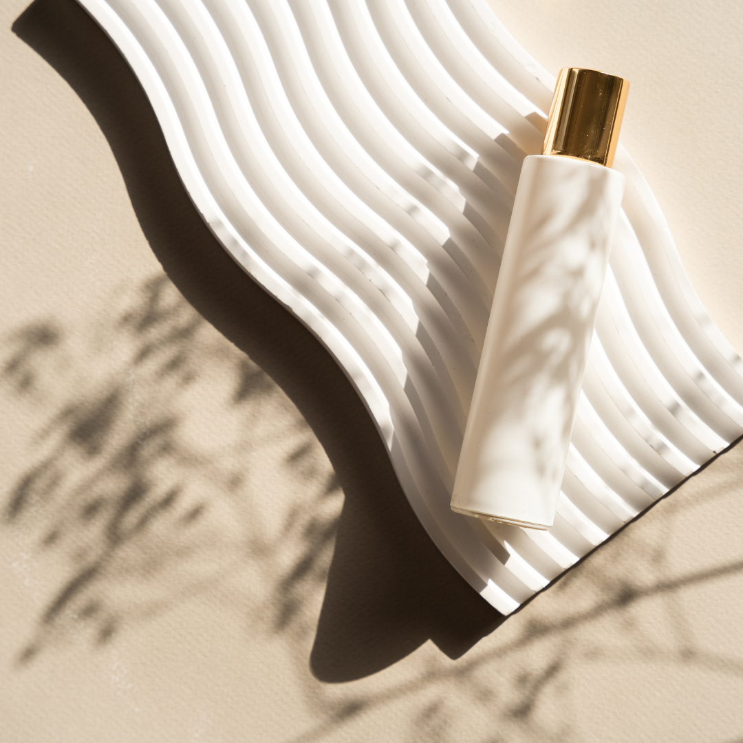 A white and gold cosmetic bottle resting on a wavy white tray, casting shadows on a beige surface.