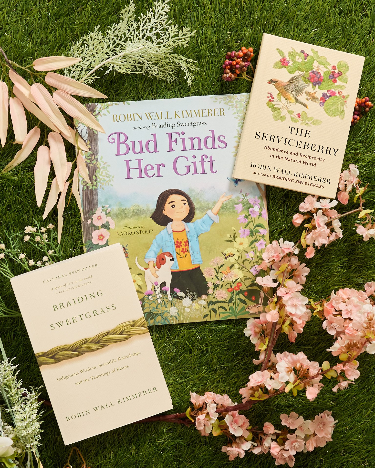A display of books and flowers on grass, including "Bud Finds Her Gift" by Robin Wall Kimmerer, "The Serviceberry" by Robin Wall Kimmerer, and "Braiding Sweetgrass" by Robin Wall Kimmerer, surrounded by pink and white blossoms and greenery.