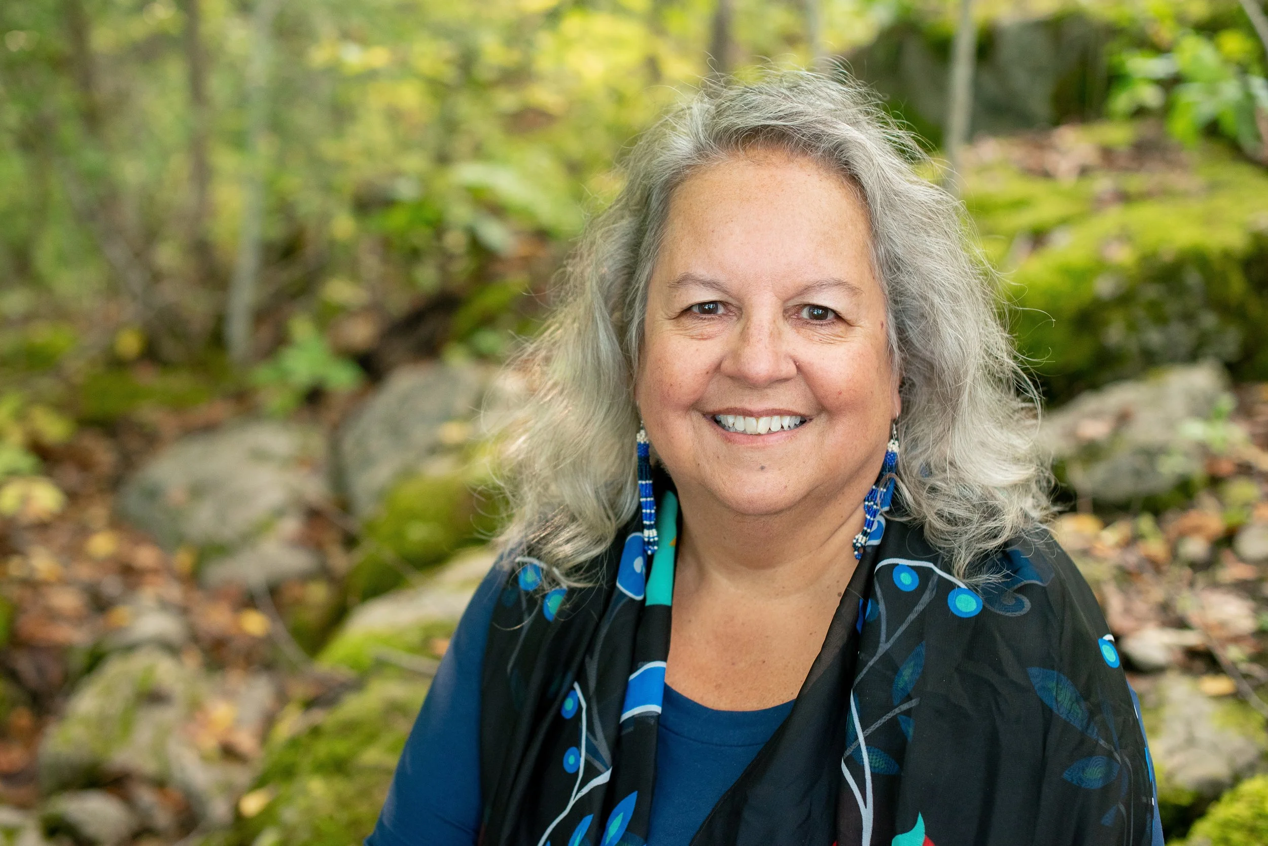 A smiling woman with gray hair and blue earrings outdoors in a forest.