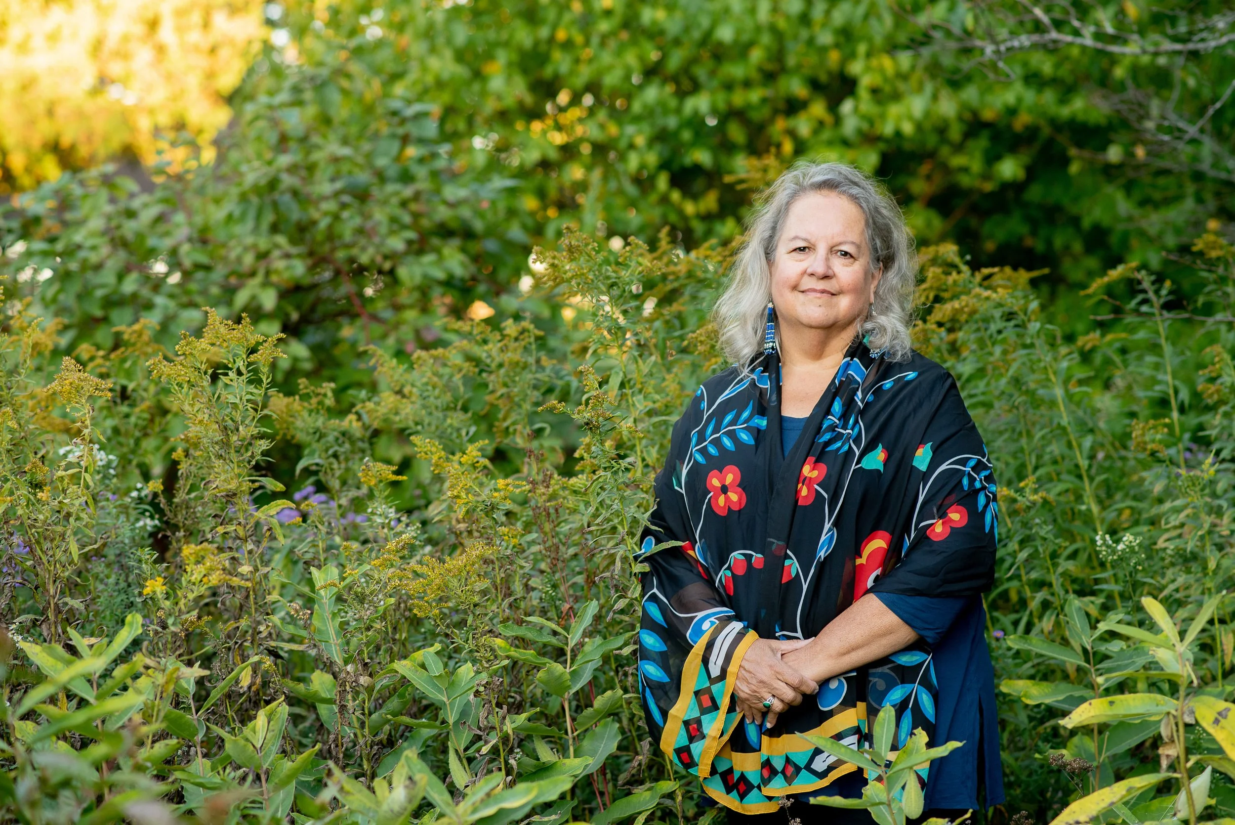 An older woman with gray hair standing outdoors in a green, leafy setting, wearing a colorful, patterned shawl with floral and geometric designs.