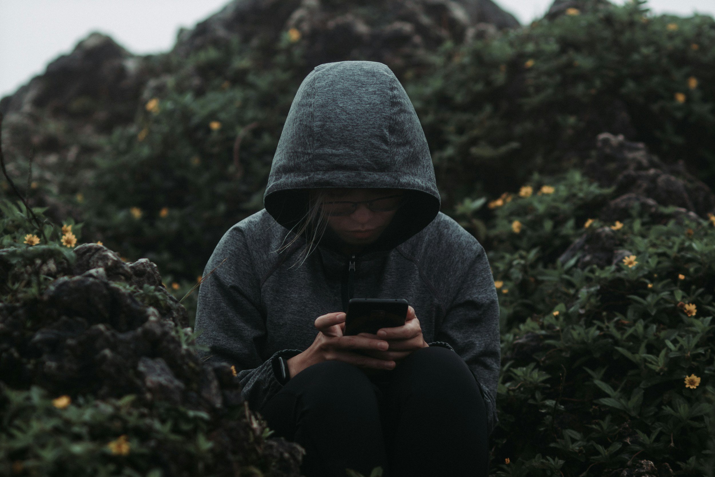 A person in a hoodie sits alone outdoors, looking down at their phone, conveying isolation, introspection, or emotional withdrawal in a quiet, natural setting.