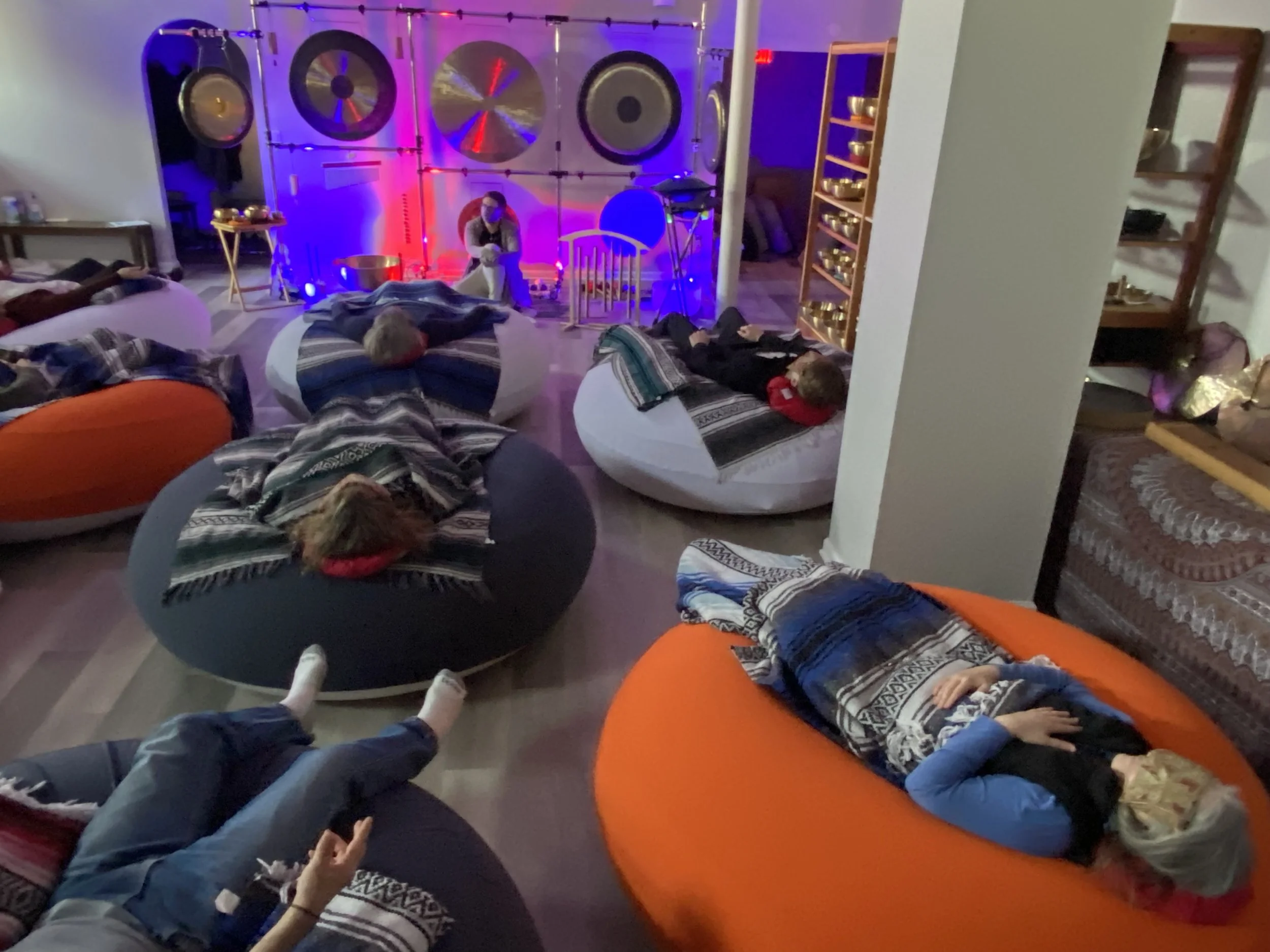 People lying on floor cushions during a sound bath session, surrounded by gongs and singing bowls lit with colorful purple and red lighting.