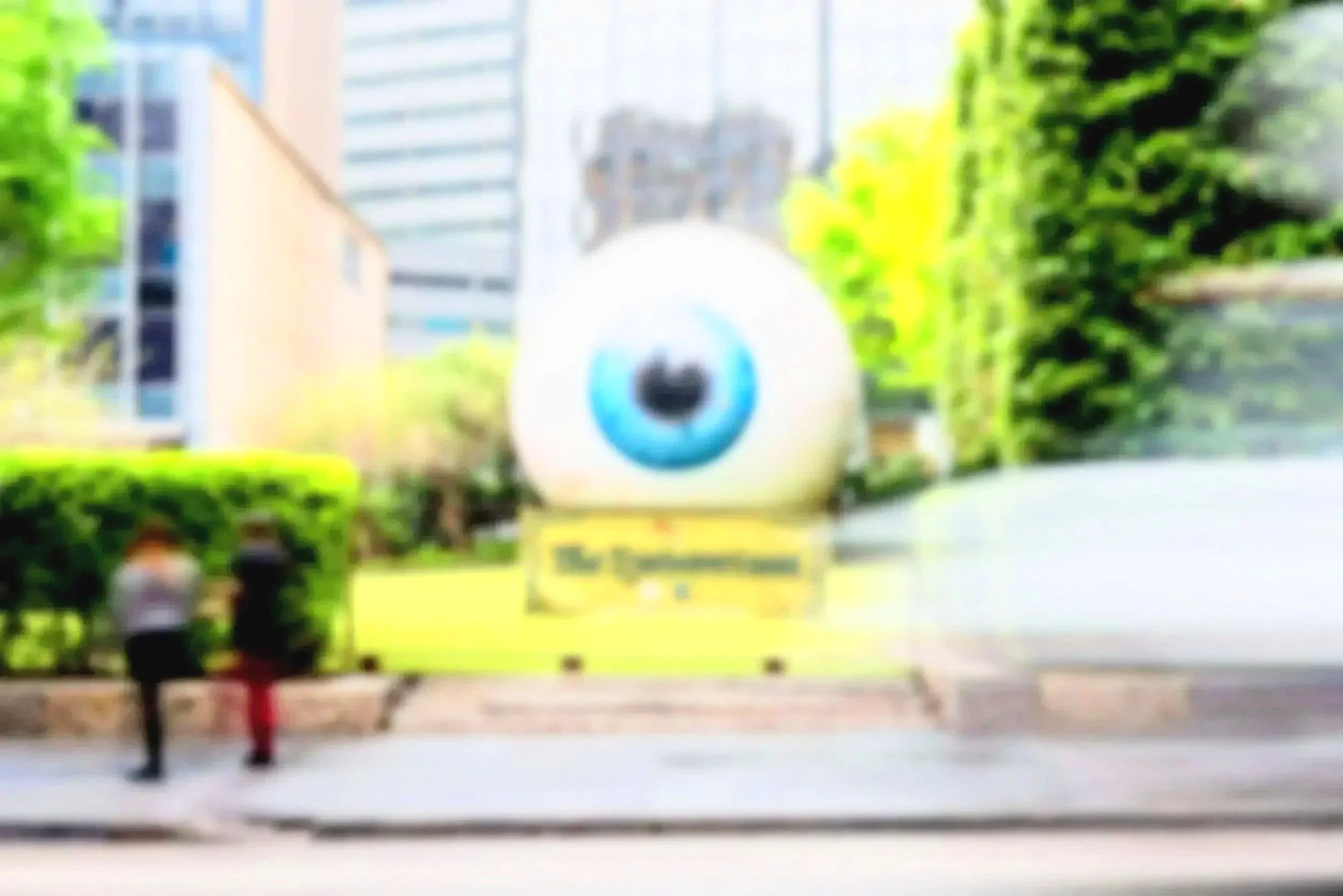 Blurred street scene with a large decorative eyeball sculpture, green trees, and tall buildings in the background.