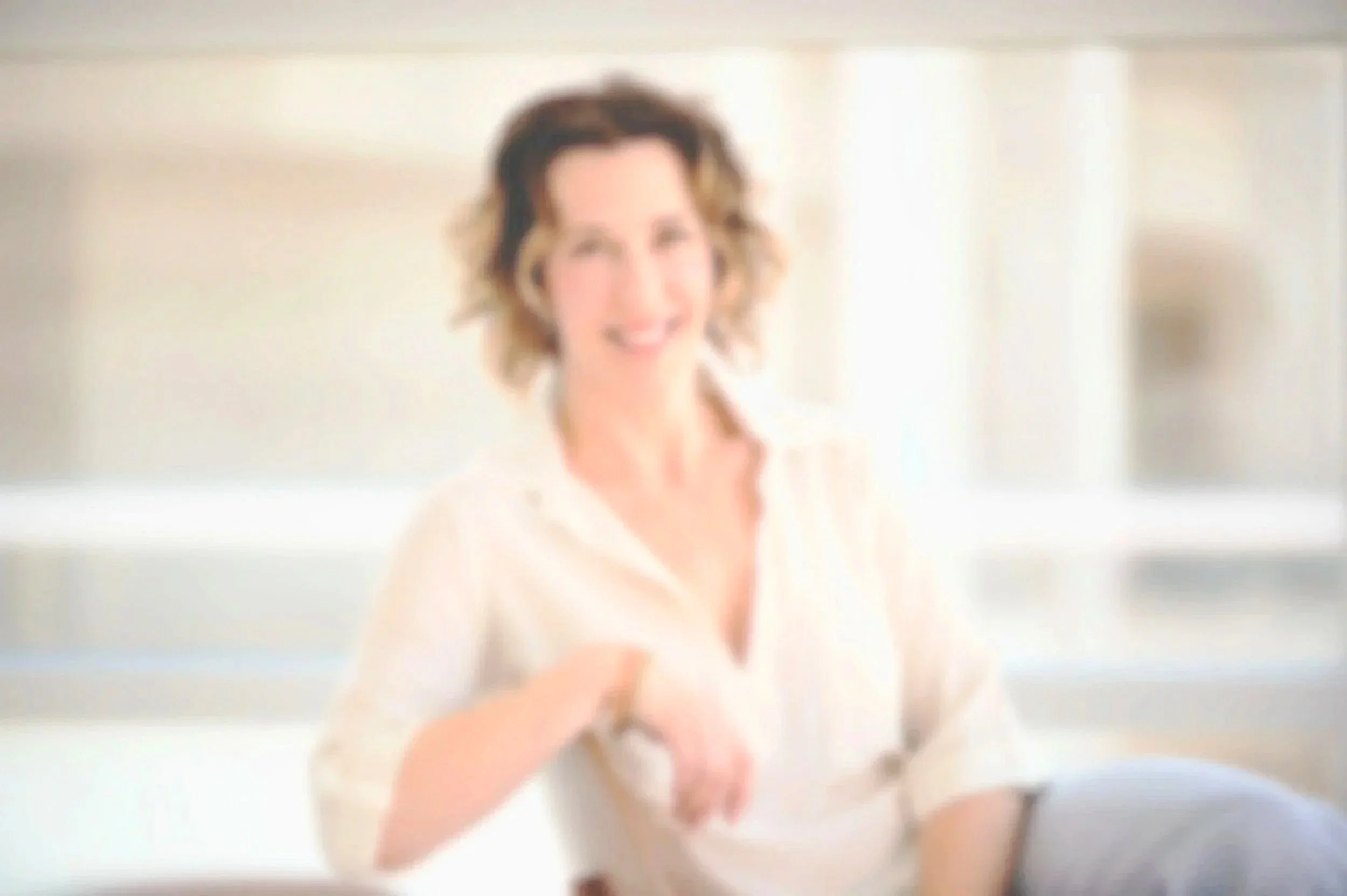 A smiling woman with short, curly hair, wearing a light-colored blouse, sitting in a bright, modern kitchen.