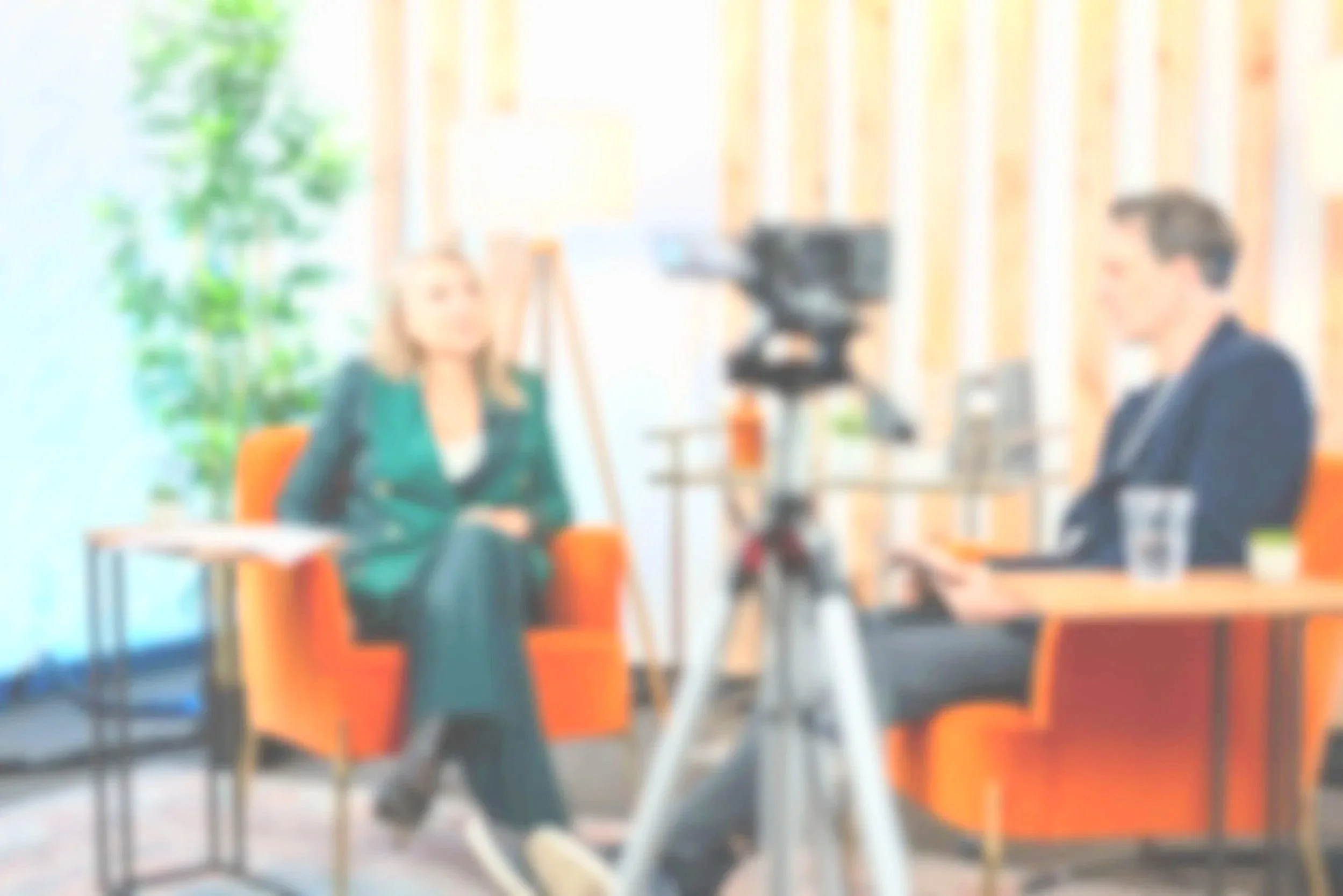 Blurred image of a woman being interviewed by a man in a studio with camera equipment and orange chairs.