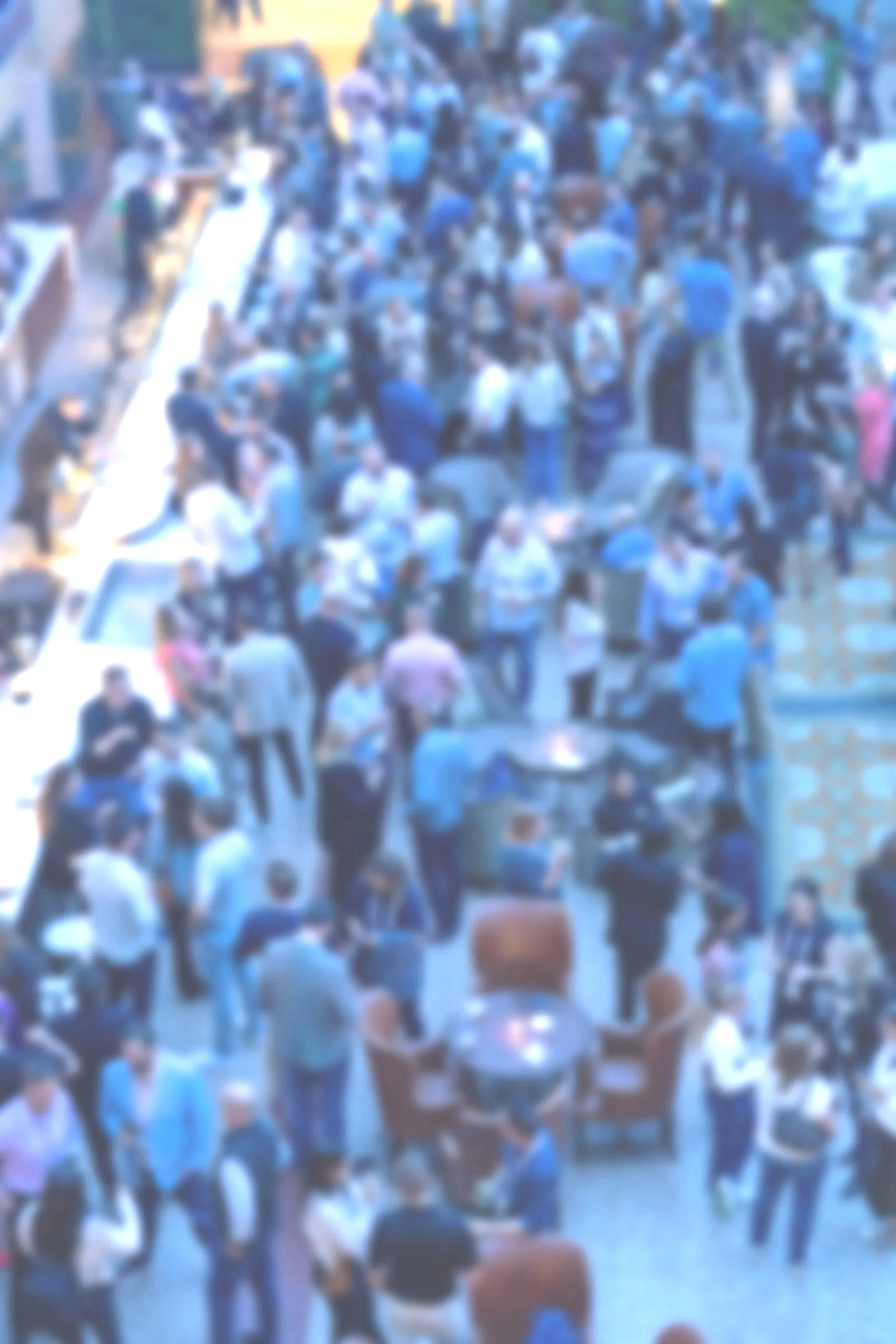 A crowded outdoor gathering with numerous people mingling, standing, and sitting around tables in a social setting.