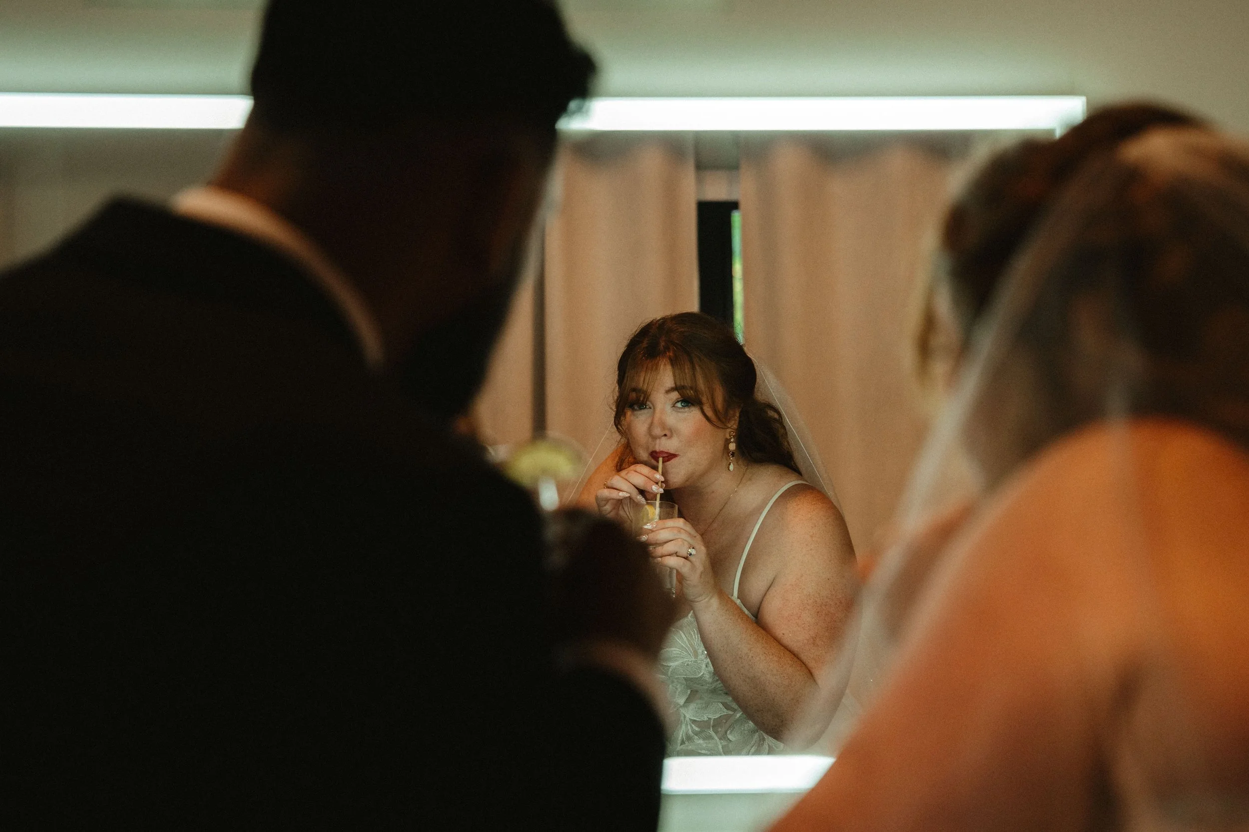 A bride with red hair and freckles, wearing a white wedding dress with thin straps, drinking from a glass straw through a mirror or reflective surface, with two people blurred in the foreground.