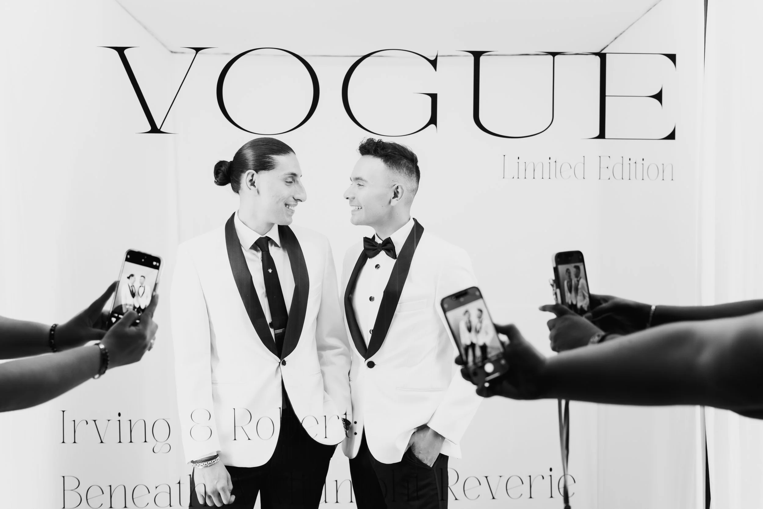 Two men in white tuxedos with black lapels, one with a bun and the other with short hair, smiling and looking at each other, surrounded by people taking photos with smartphones at a Vogue Limited Edition event.