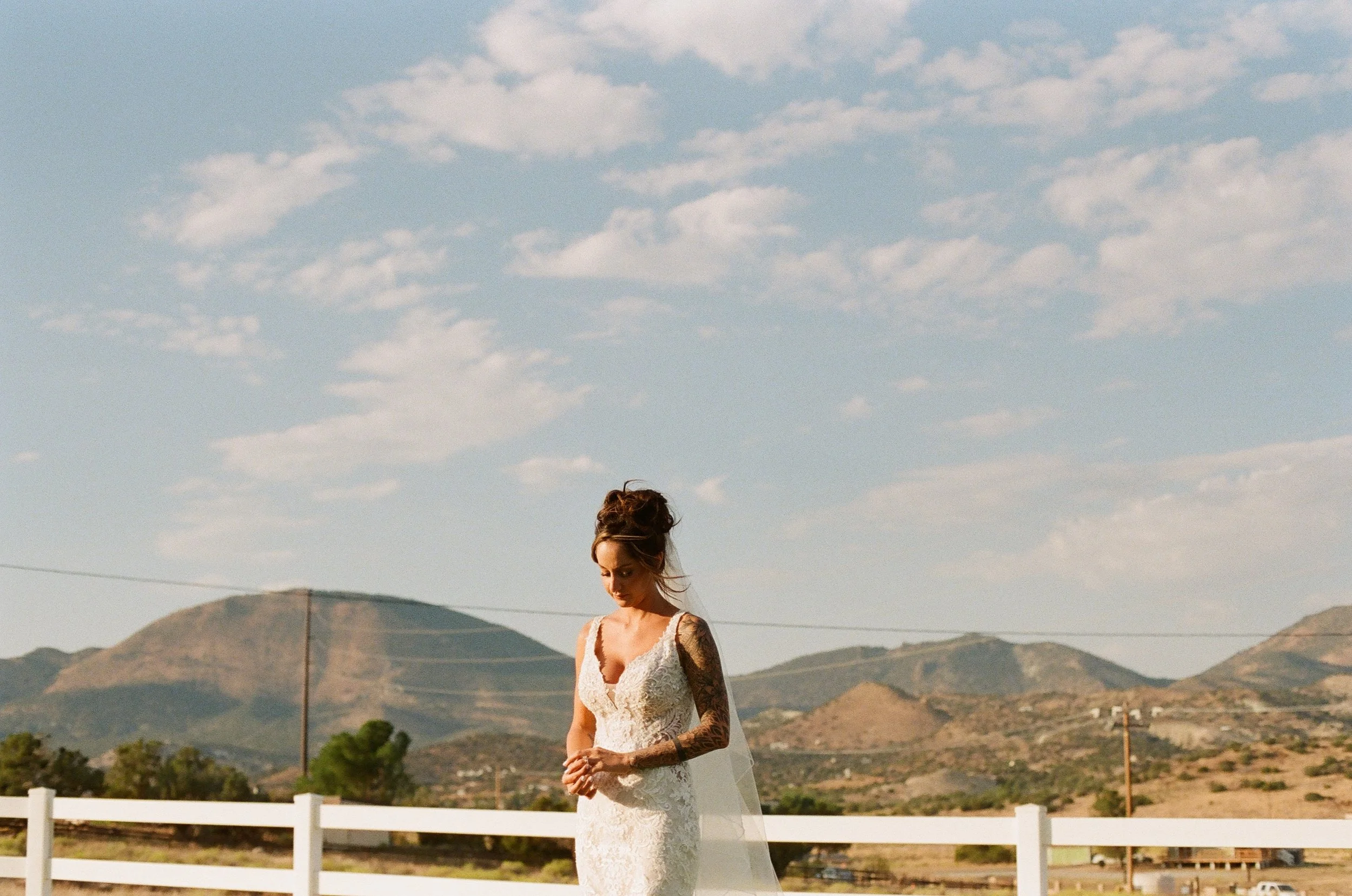 A woman in a white lace wedding dress standing outdoors with mountains in the background, looking down with her hair styled in an elegant updo.