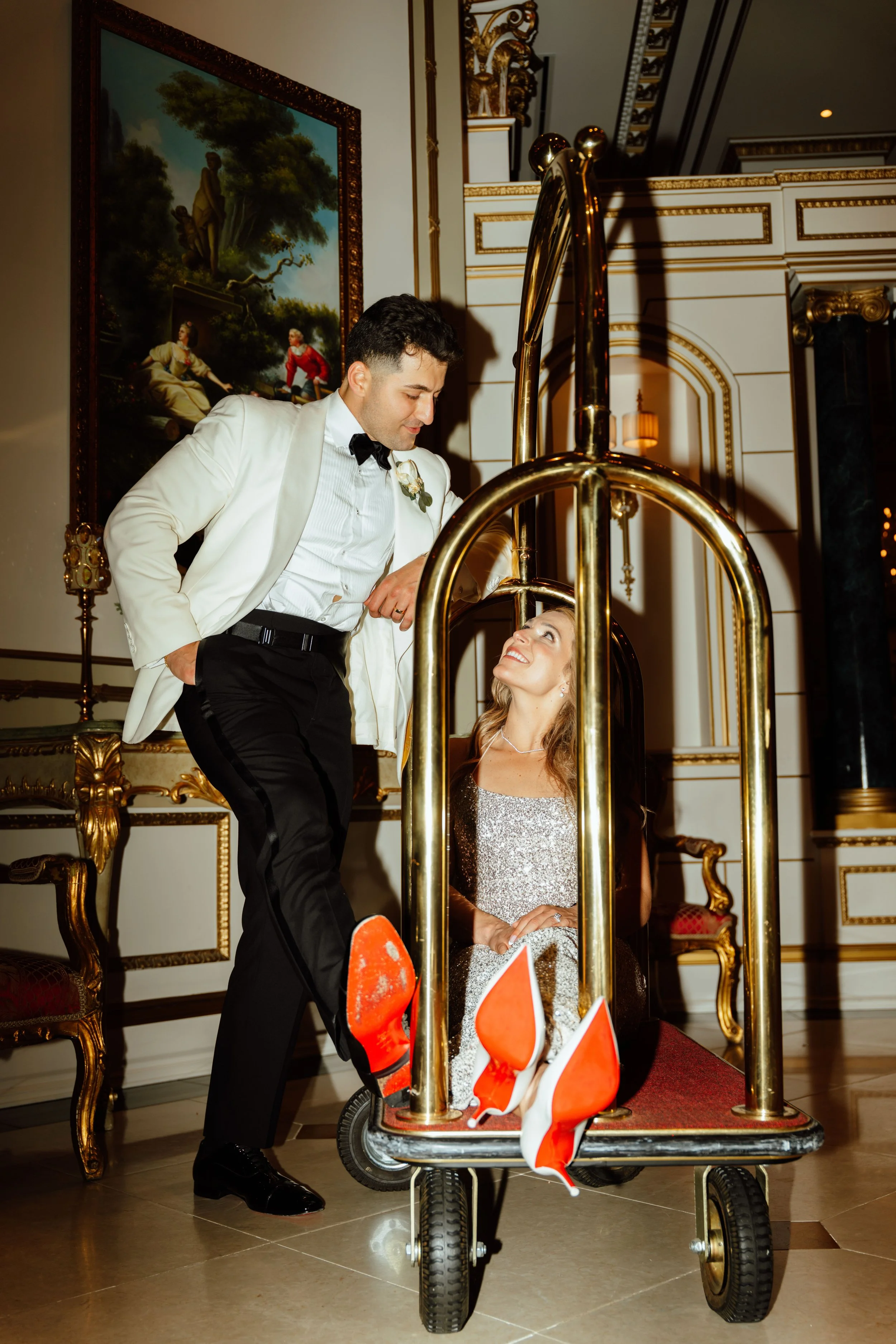 A man in a white tuxedo with a black bow tie, standing next to a woman seated in a luxury hotel lobby. The woman is wearing a sparkly silver dress, smiling, and looking at the man. They are beside a luggage cart with bright red high-heeled shoes on t