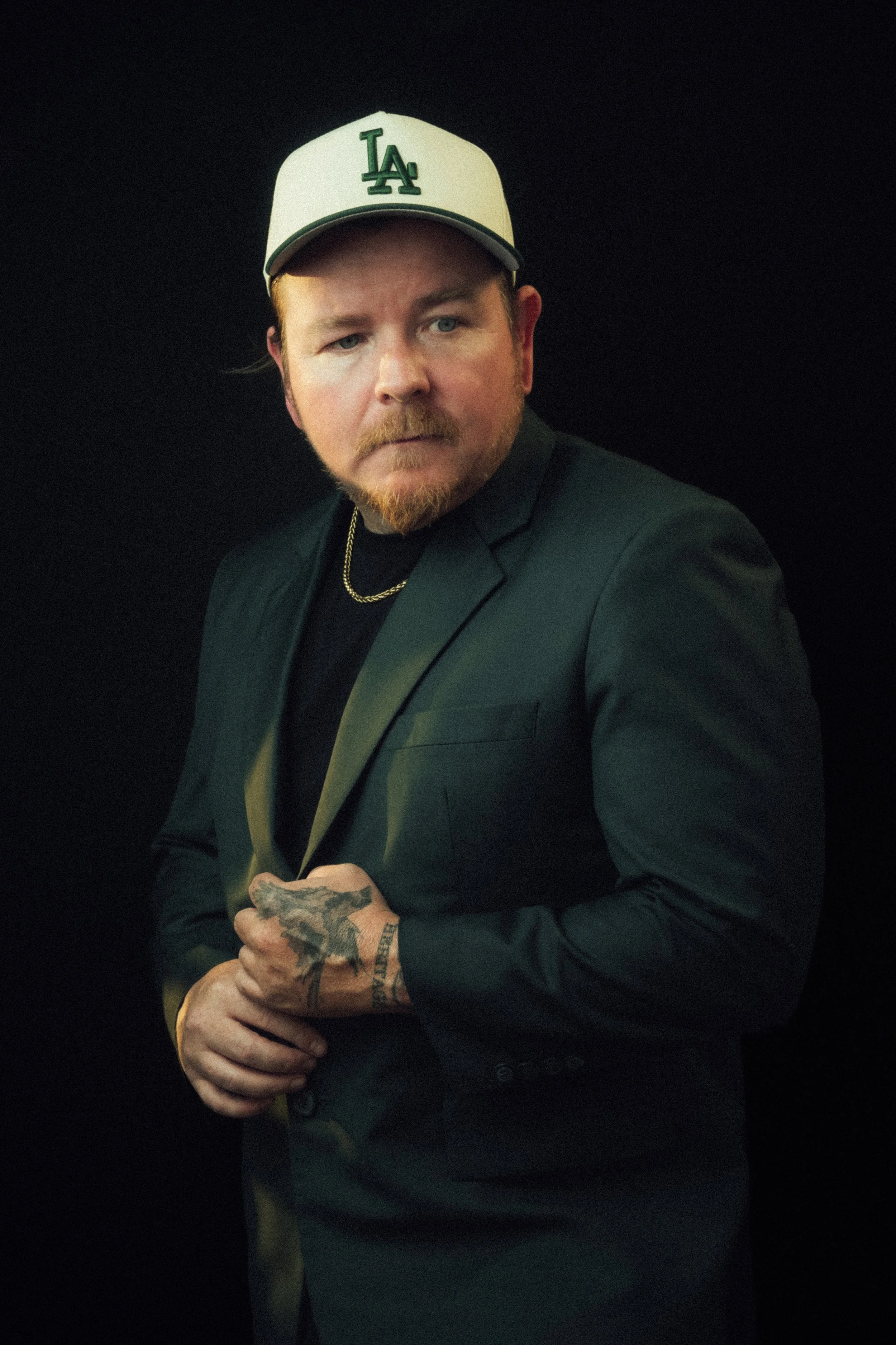 A man with a reddish beard and mustache wearing a white LA Dodgers baseball cap, a black blazer, a chain necklace, and tattooed hands, posing against a black background.