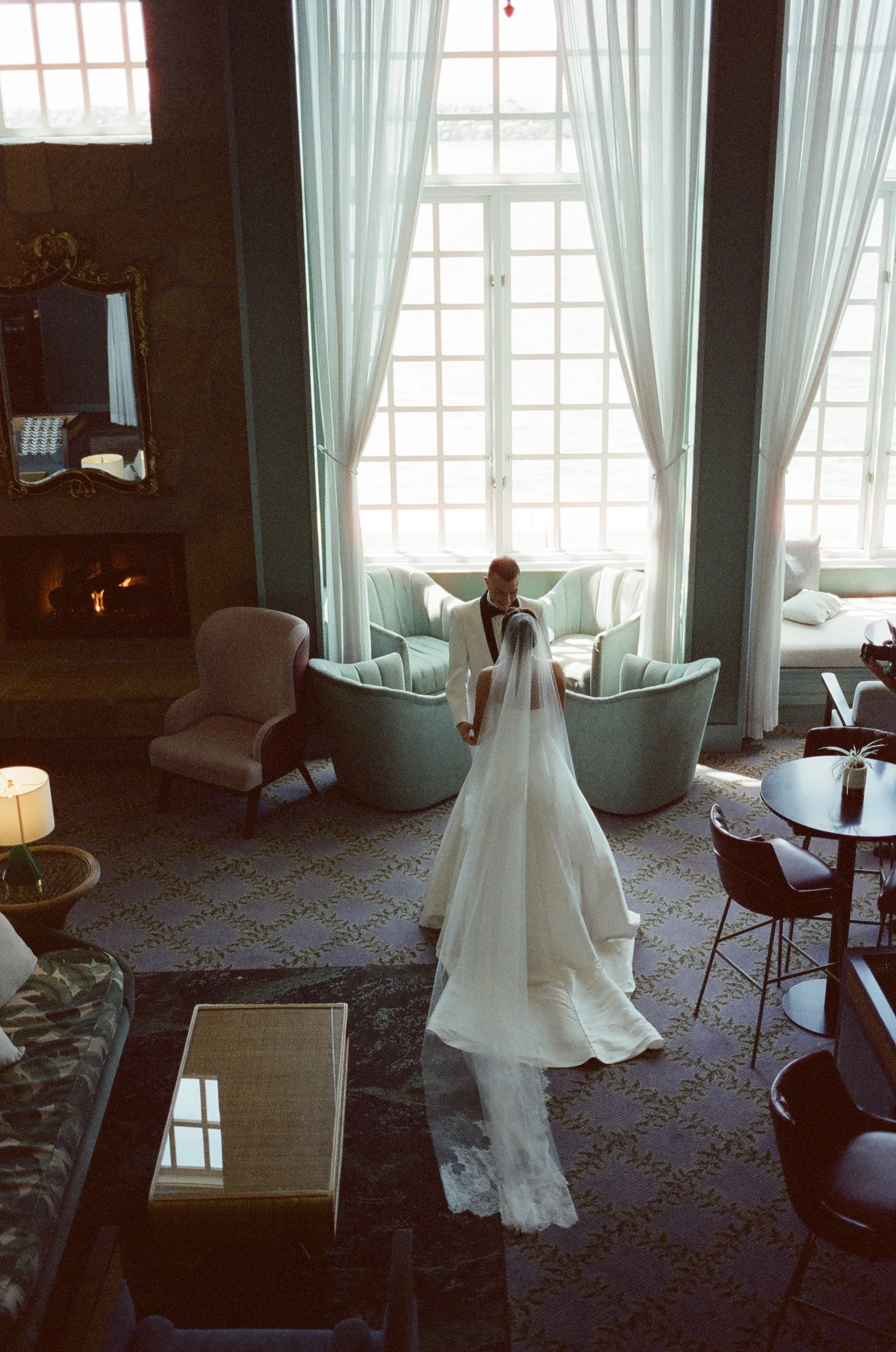 A bride and groom stand inside a large, bright room with tall windows and white curtains. The bride wears a long, white wedding dress with a veil, and the groom is dressed in a white tuxedo.