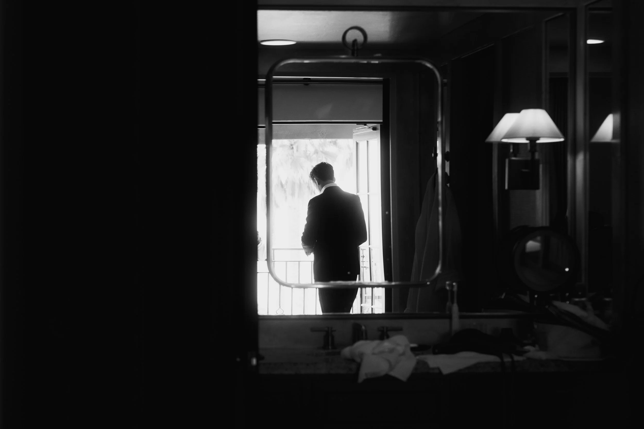 A person in a suit standing by a window in a room, viewed through a mirror in a black and white photo.
