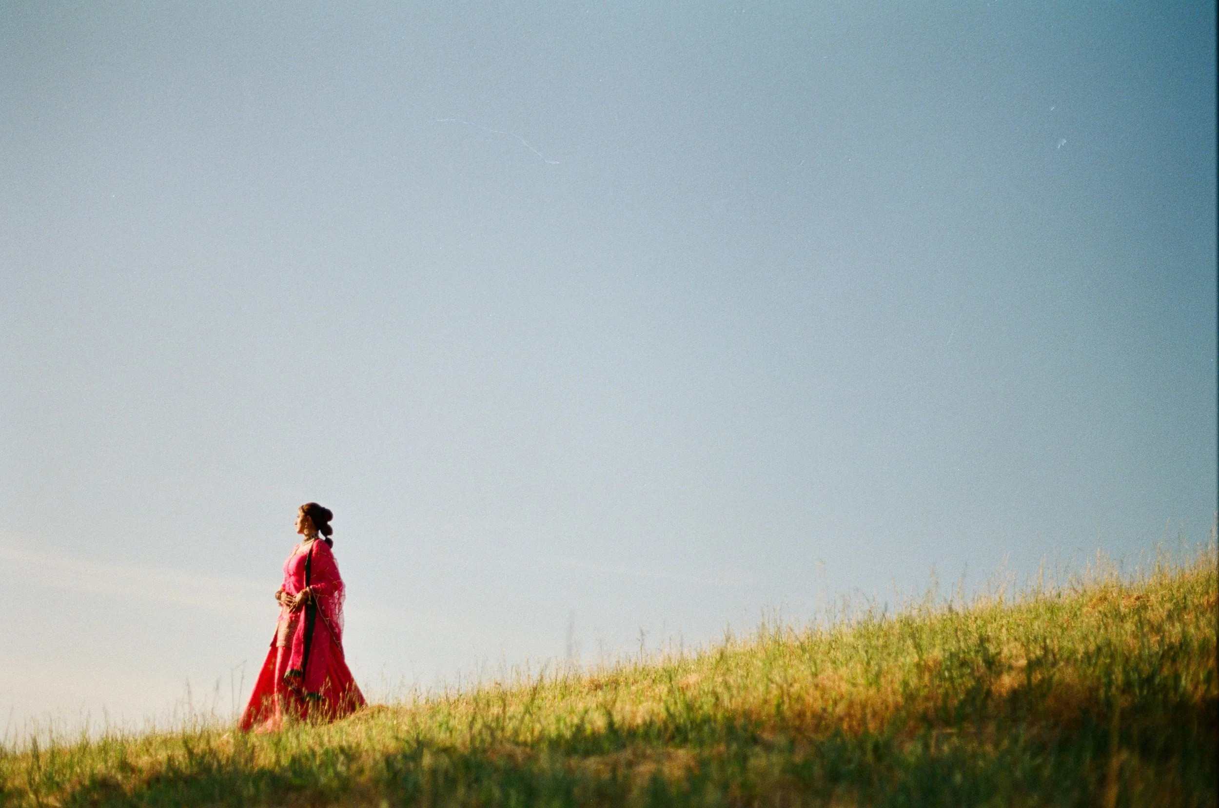 A woman in a traditional pink and red dress walking on a grassy hill under a clear blue sky.