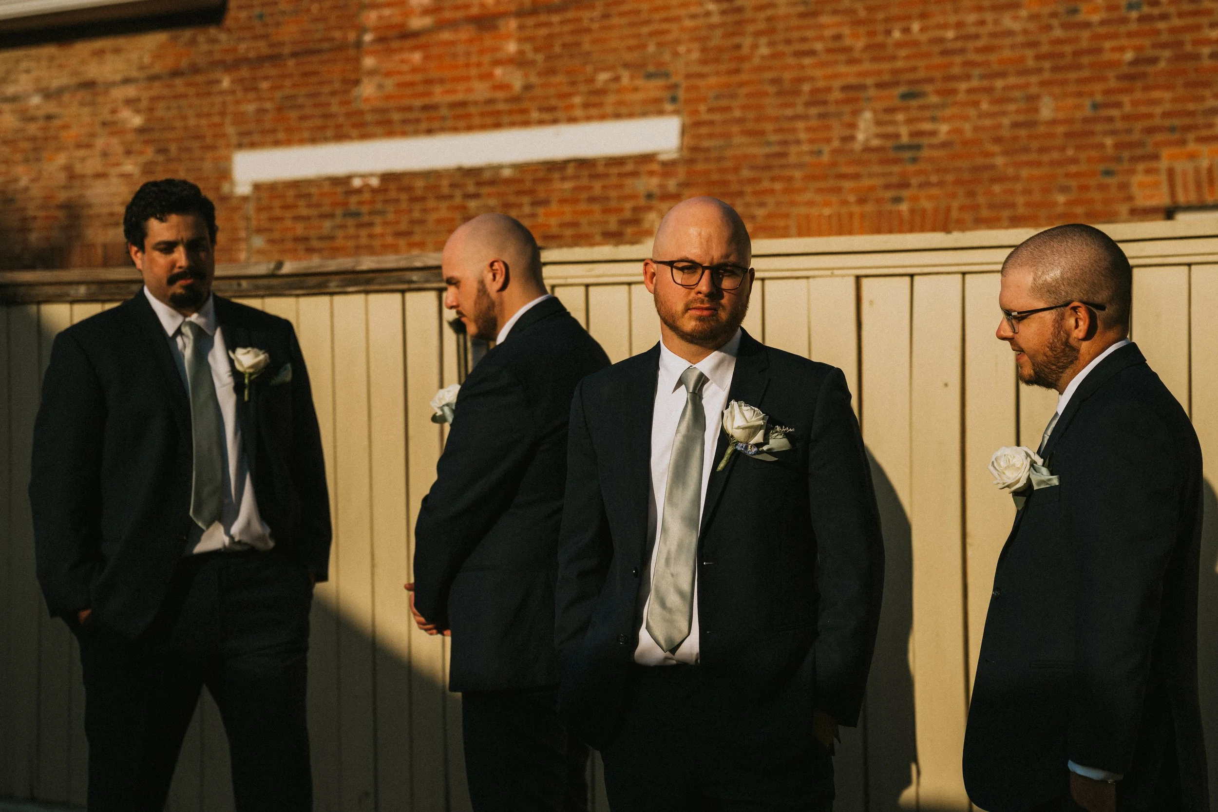 Four men in suits and glasses standing outdoors at night against a brick wall and wooden fence.