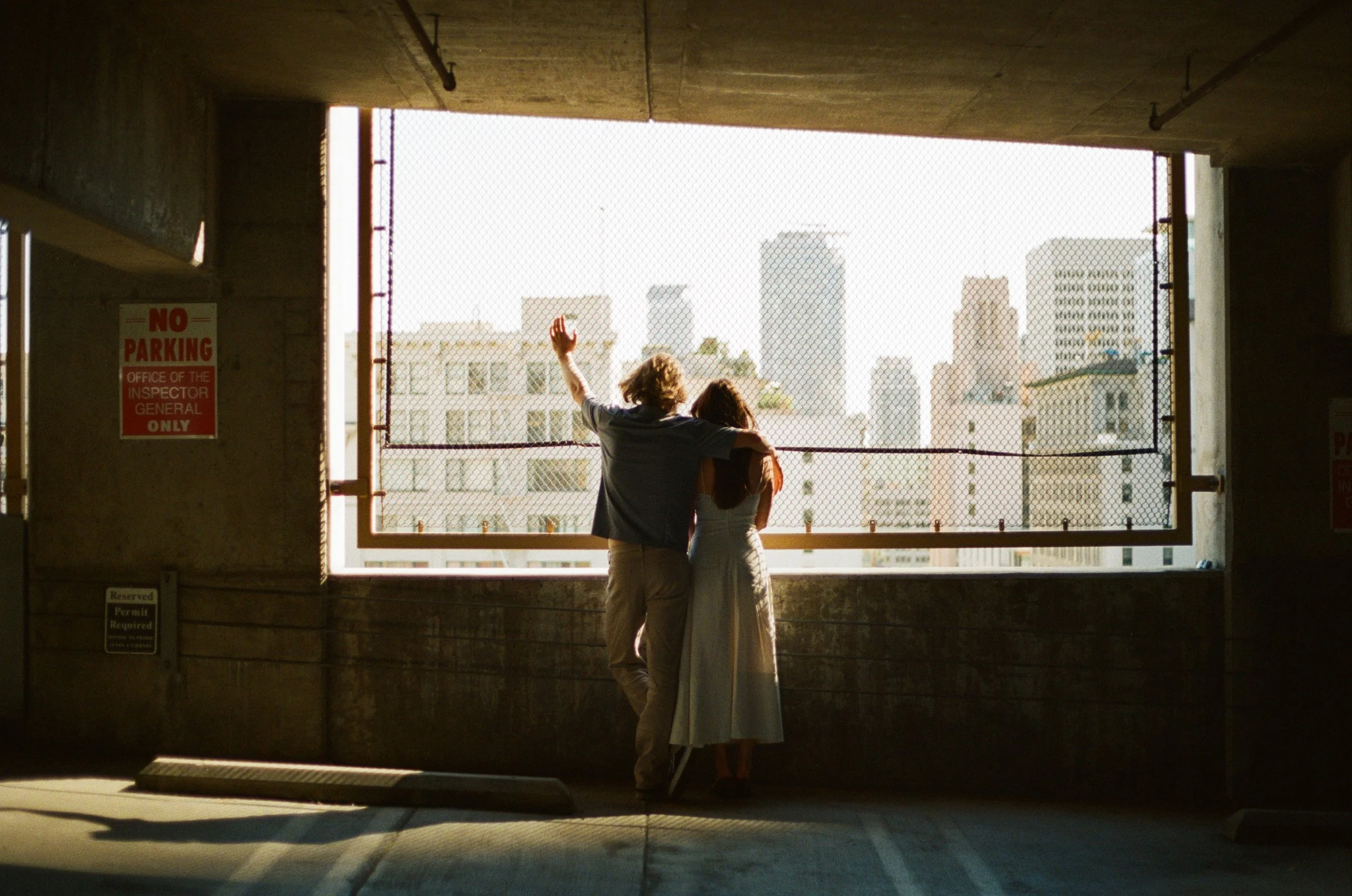 Two people standing on a balcony, overlooking a city skyline through a chain-link fence. One person is waving.