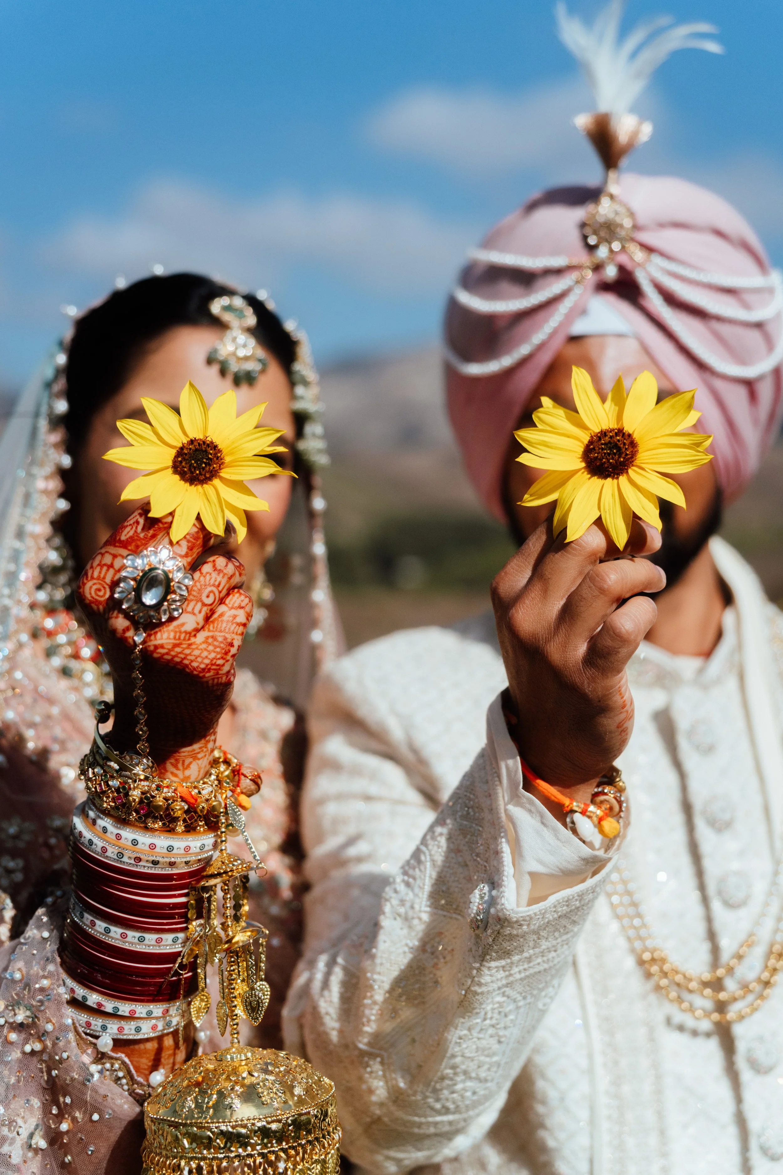 A man and a woman dressed in traditional Indian wedding attire holding yellow sunflowers, with jewelry and a cloudy blue sky in the background.