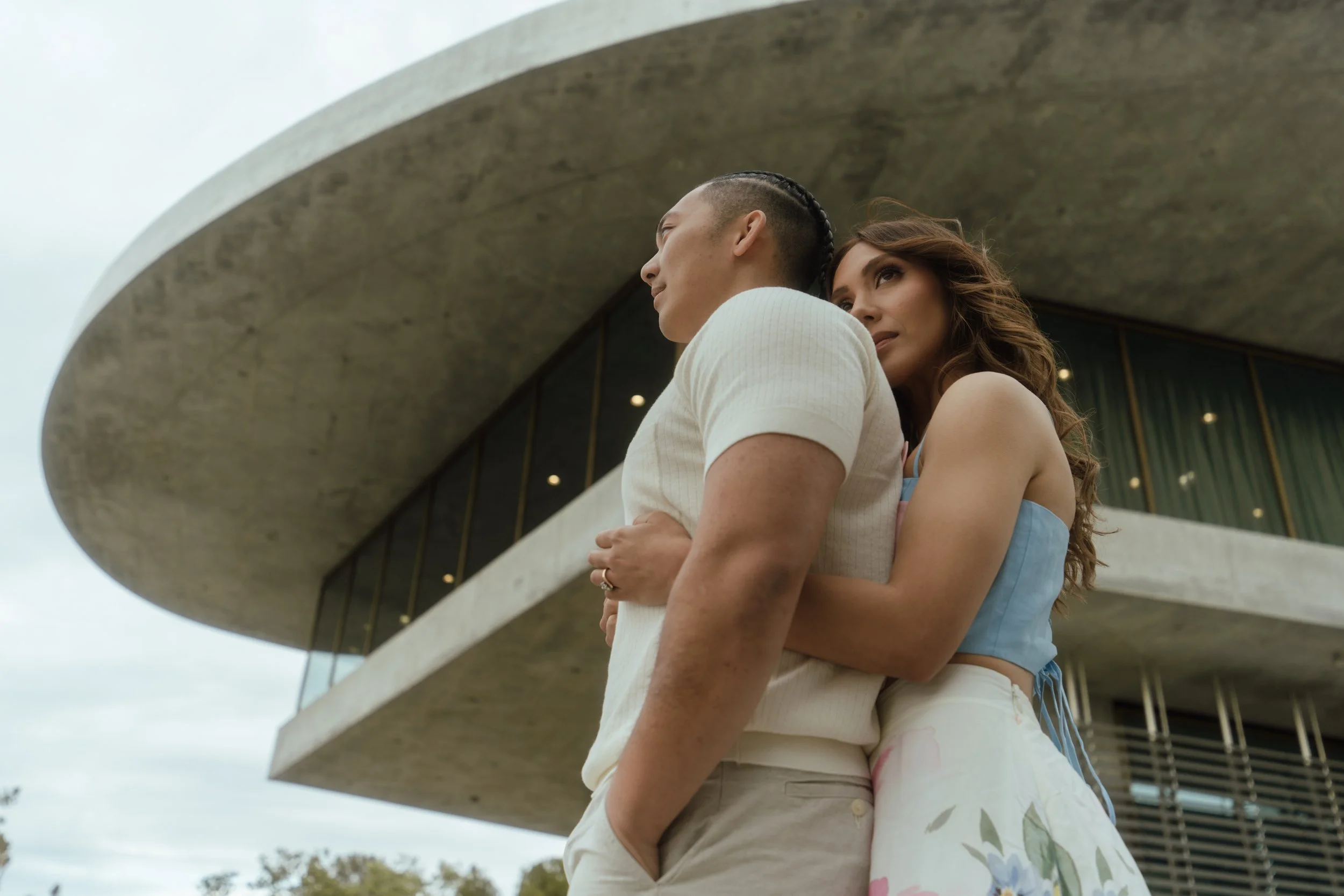 A man and woman embrace each other outdoors near modern architecture with curved concrete and large glass windows, with trees in the background.