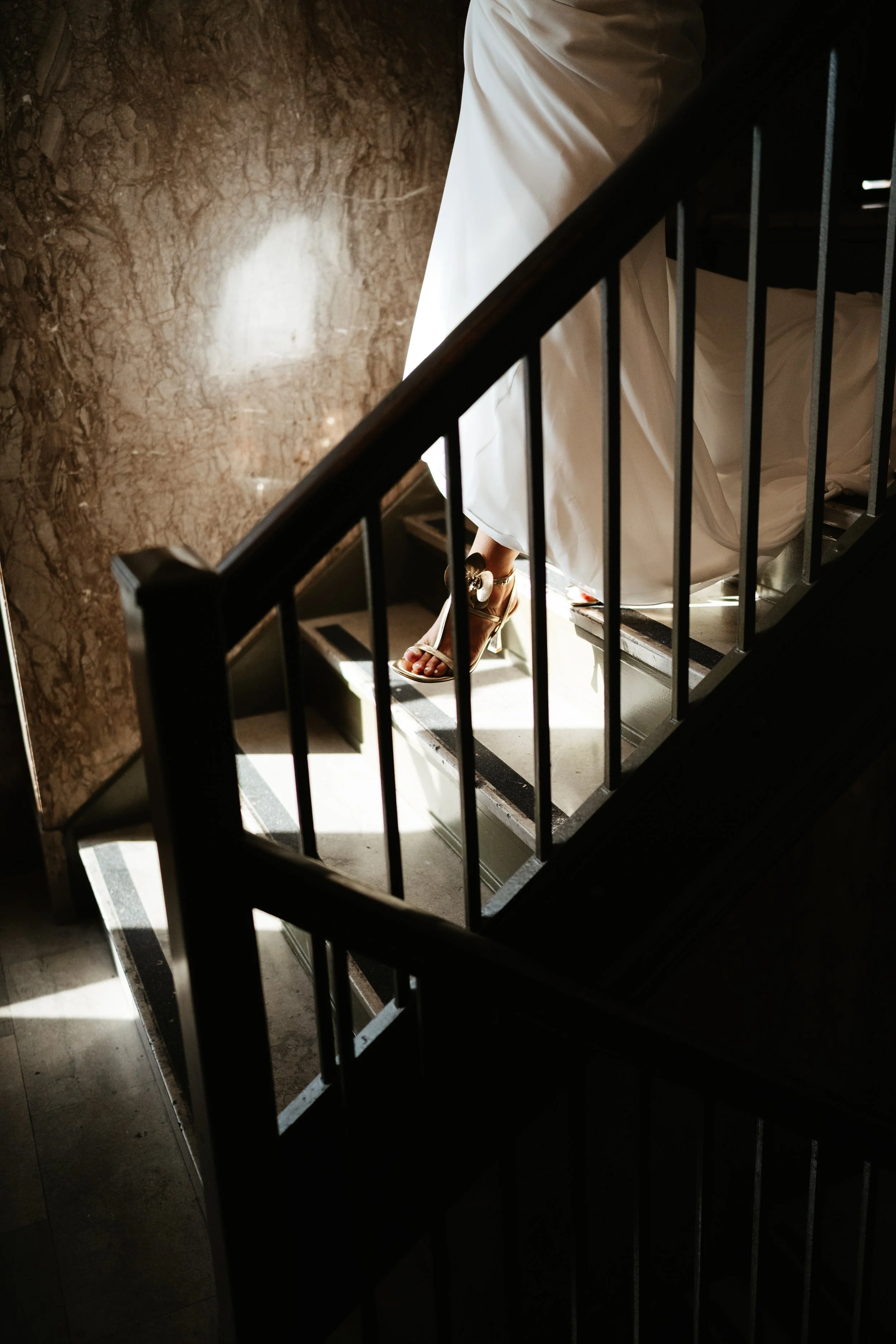 A person wearing a white dress and high-heeled sandals is descending a staircase with a black railing. The staircase has black and white checkered flooring, and a marble wall is visible on the left side of the image, with sunlight streaming in and cr
