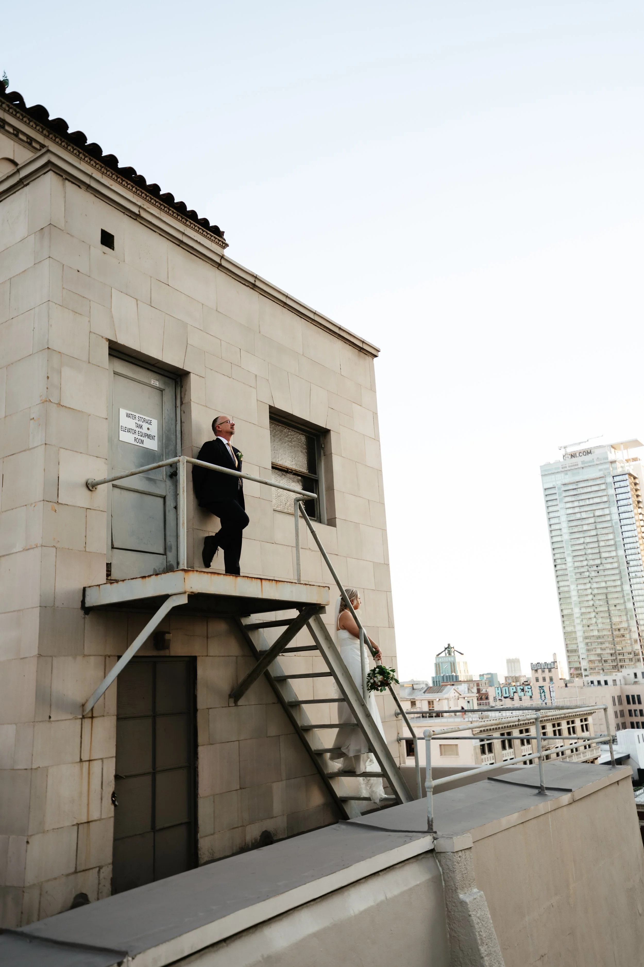 A man in a black tuxedo and a woman in a white dress on a rooftop, with the woman holding a bouquet of flowers, in an urban setting with buildings in the background.
