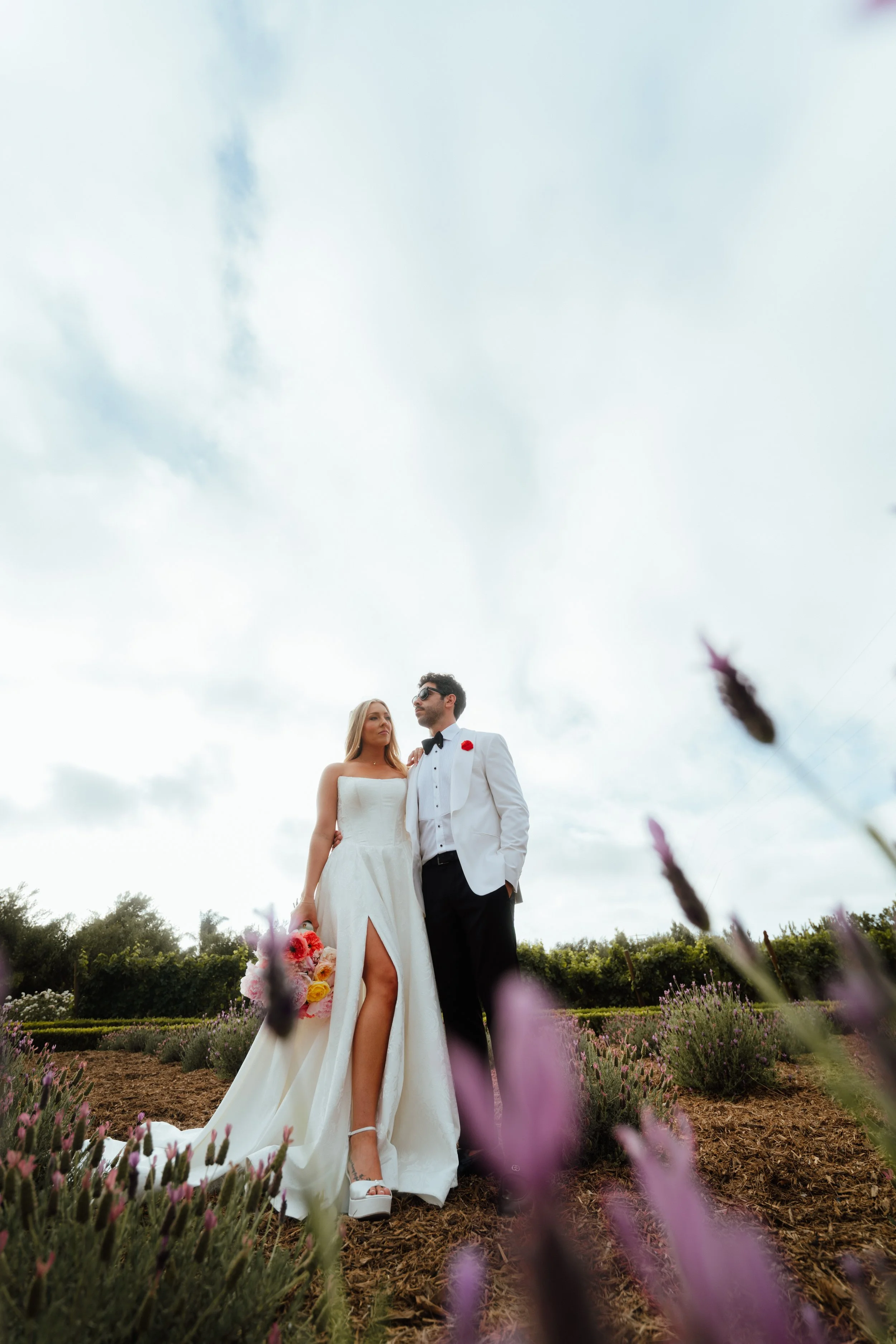 A bride and groom standing together outdoors in a garden, surrounded by pink and purple flowers, under a cloudy sky.