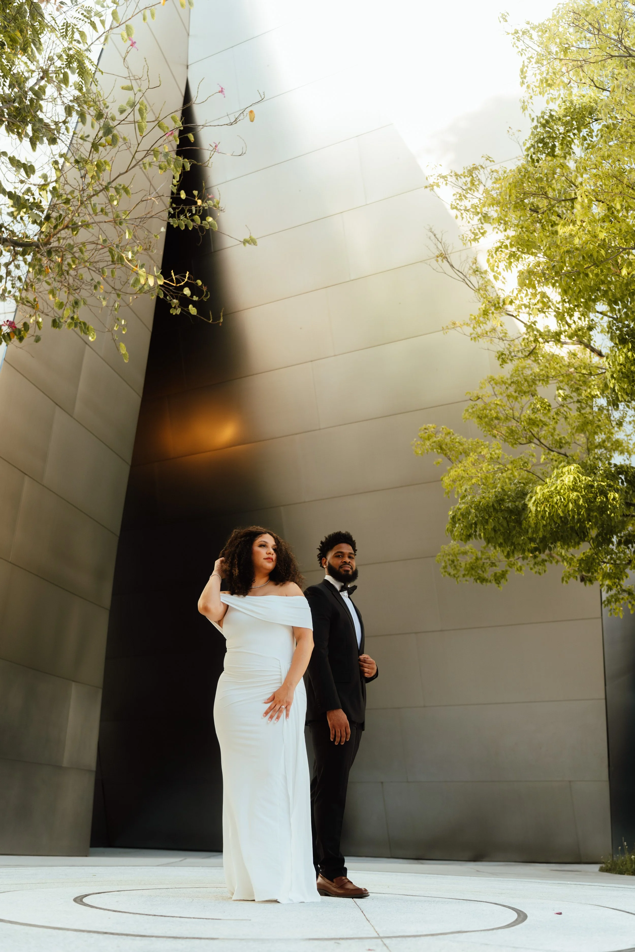 A man and woman dressed in formal attire standing on a modern outdoor stage with a curved metal wall and trees.