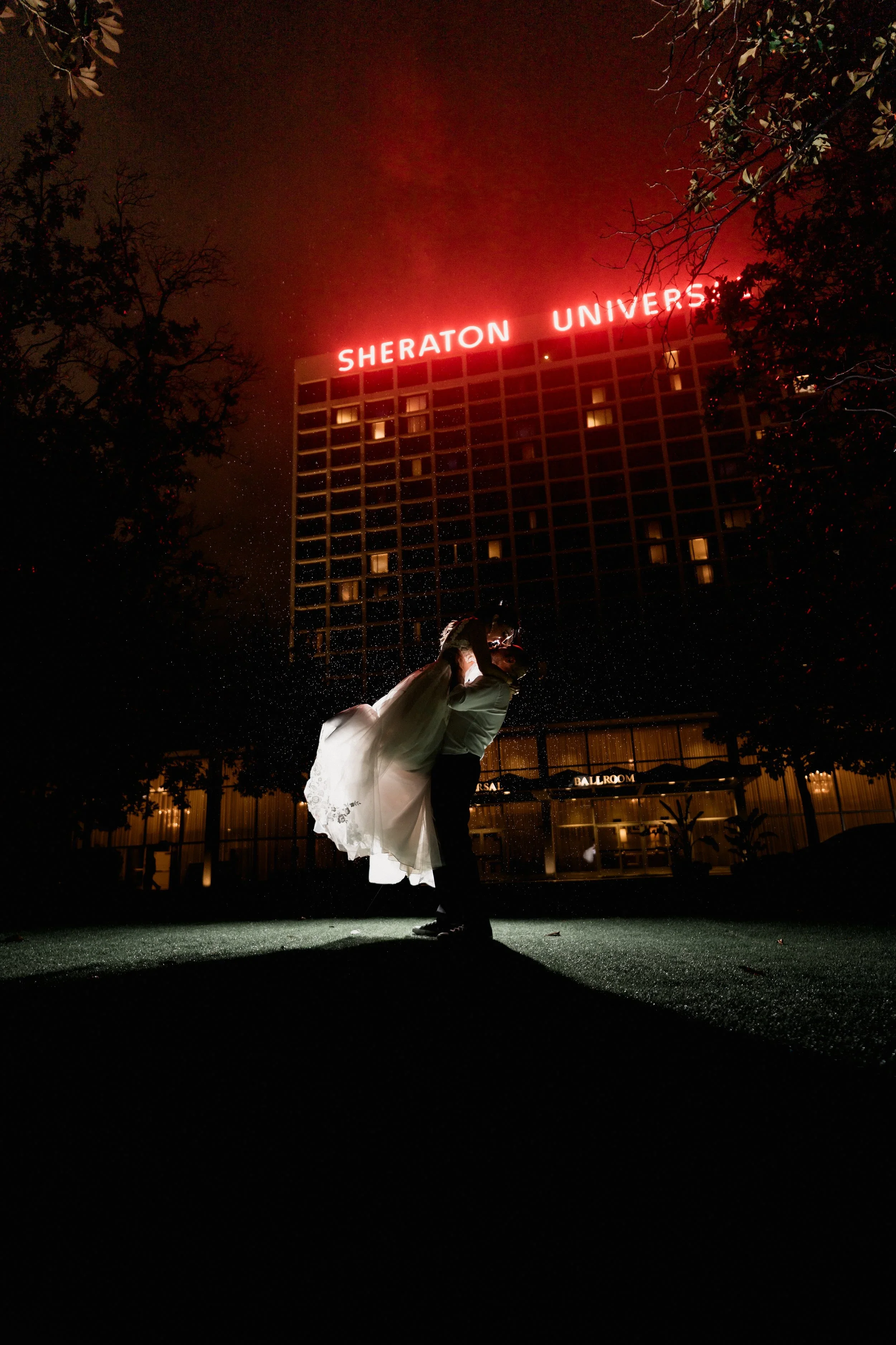 A bride and groom sharing a dance outdoors at night, backlit with darkness and city lights, with neon hotel signs in the background.