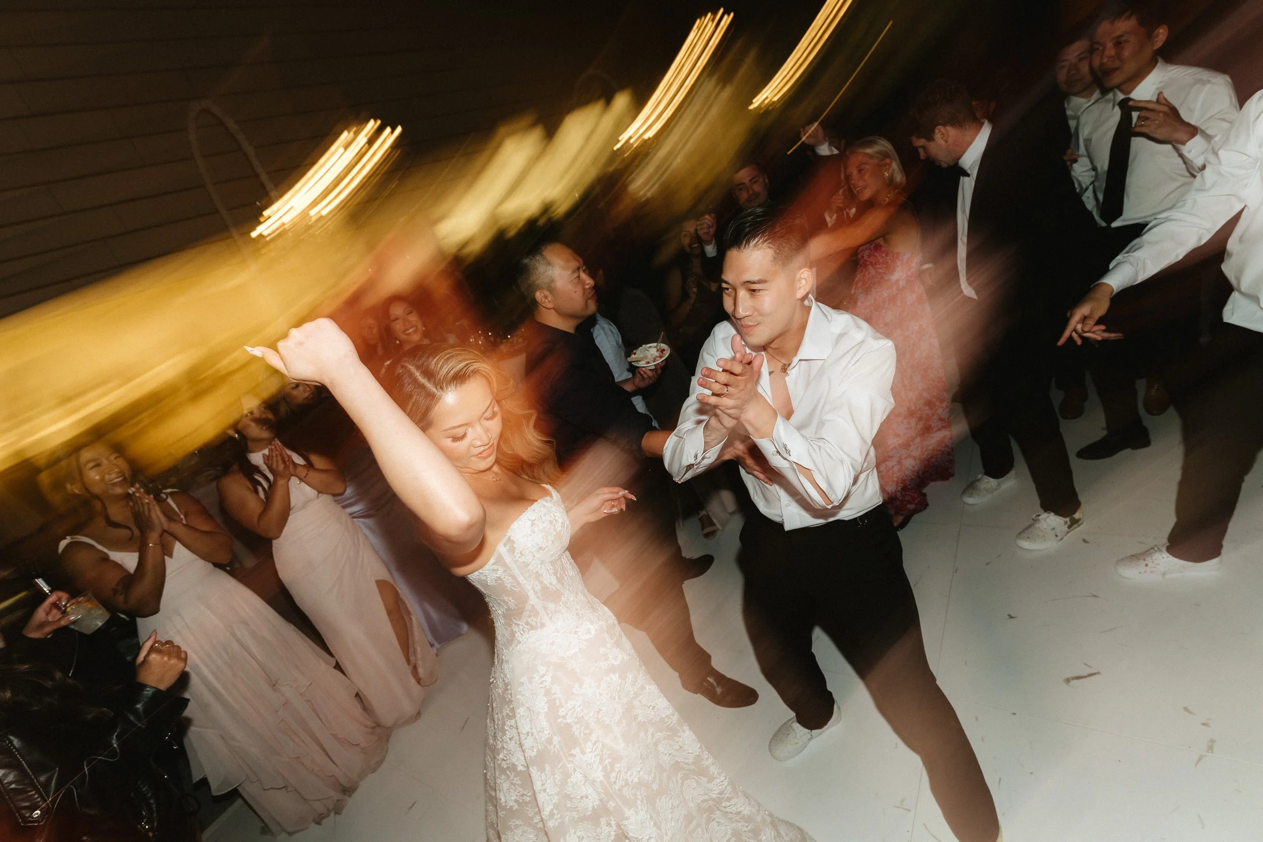 People dancing at a wedding reception, including a bride in a white lace wedding dress, with guests enjoying the celebration.